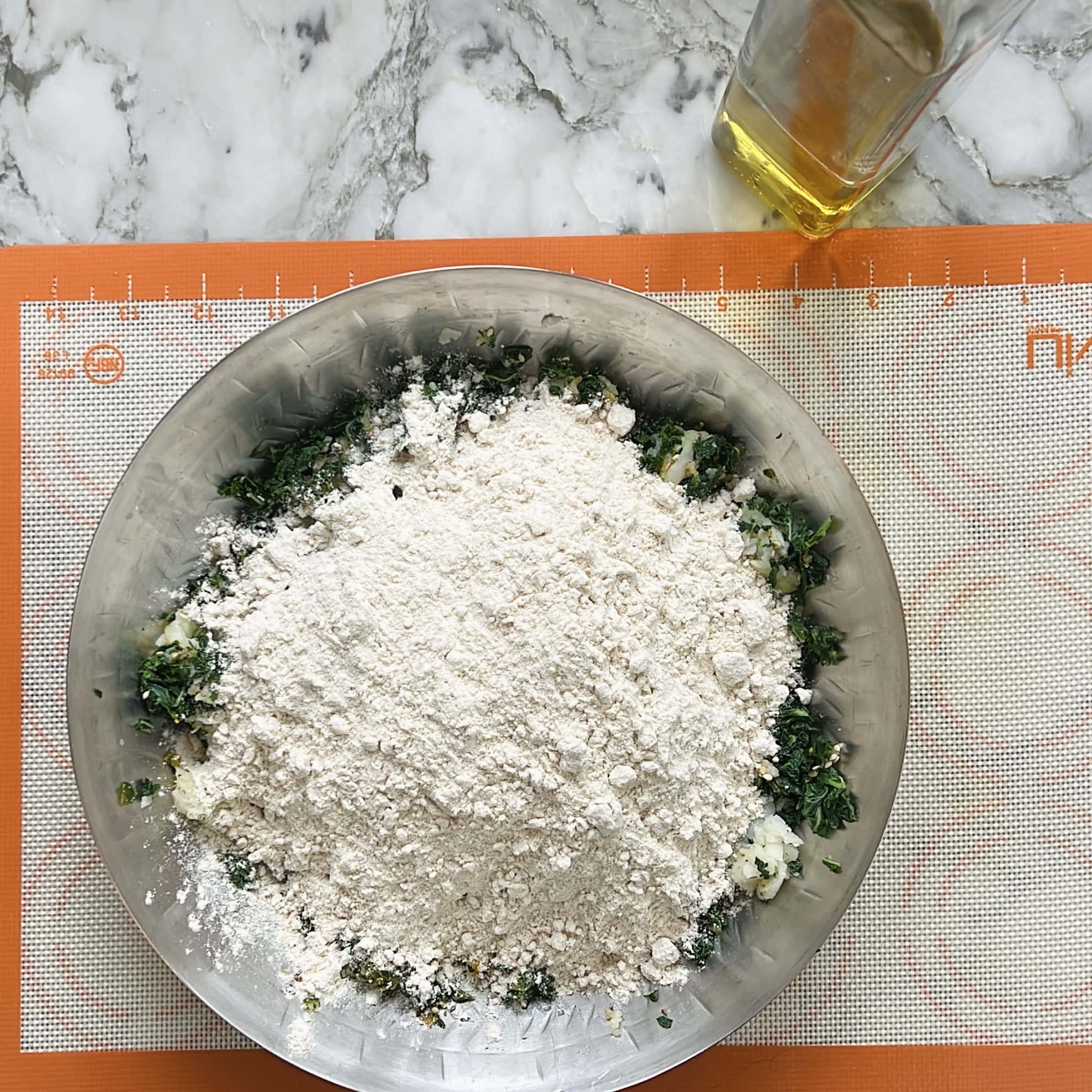 A stainless steel bowl with flour and chopped greens on a silicone baking mat, next to a bottle of oil on a marble countertop.