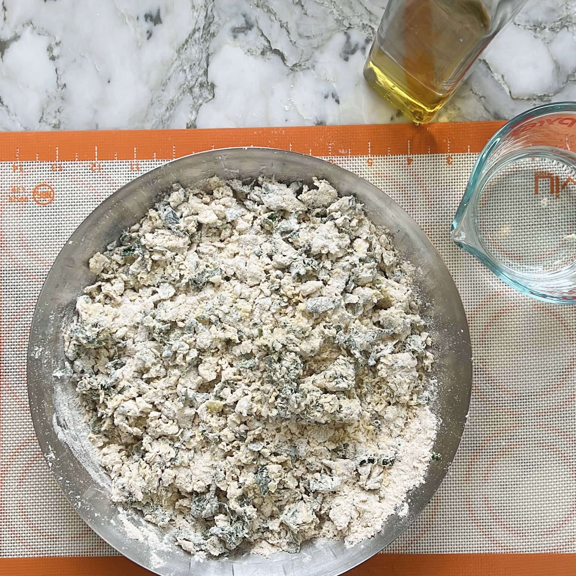 A metal bowl filled with a crumbly flour and vegetanle mixture sits on a silicone baking mat. A glass measuring cup with water and a bottle of oil are nearby on a marble counter.