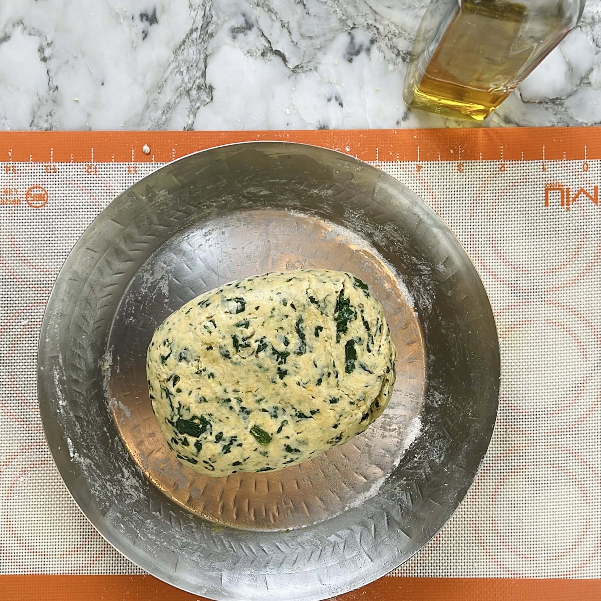 A ball of dough with kale in a metal bowl on a silicone baking mat, with a bottle of olive oil nearby on a marble countertop.