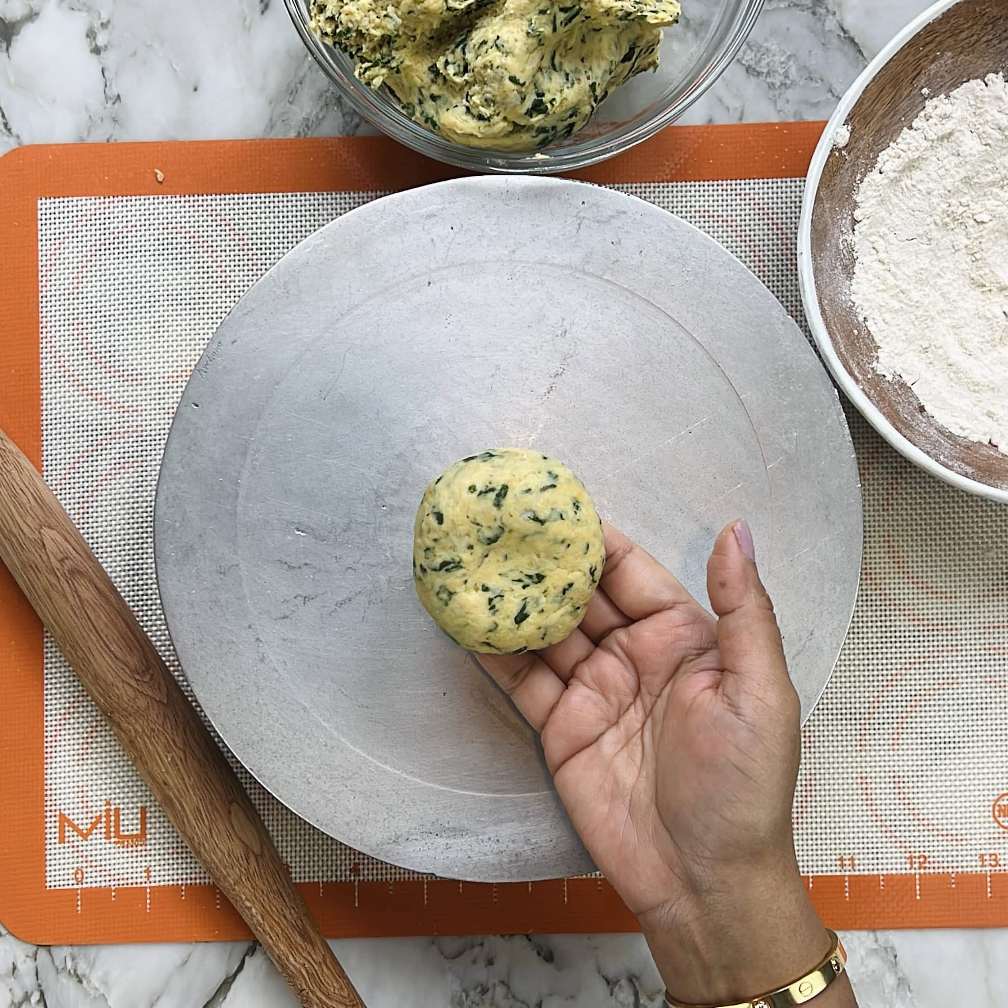 A hand holds a ball of dough with herbs over a floured metal surface, surrounded by a rolling pin, flour bowl, and bowl of dough on a silicone mat.