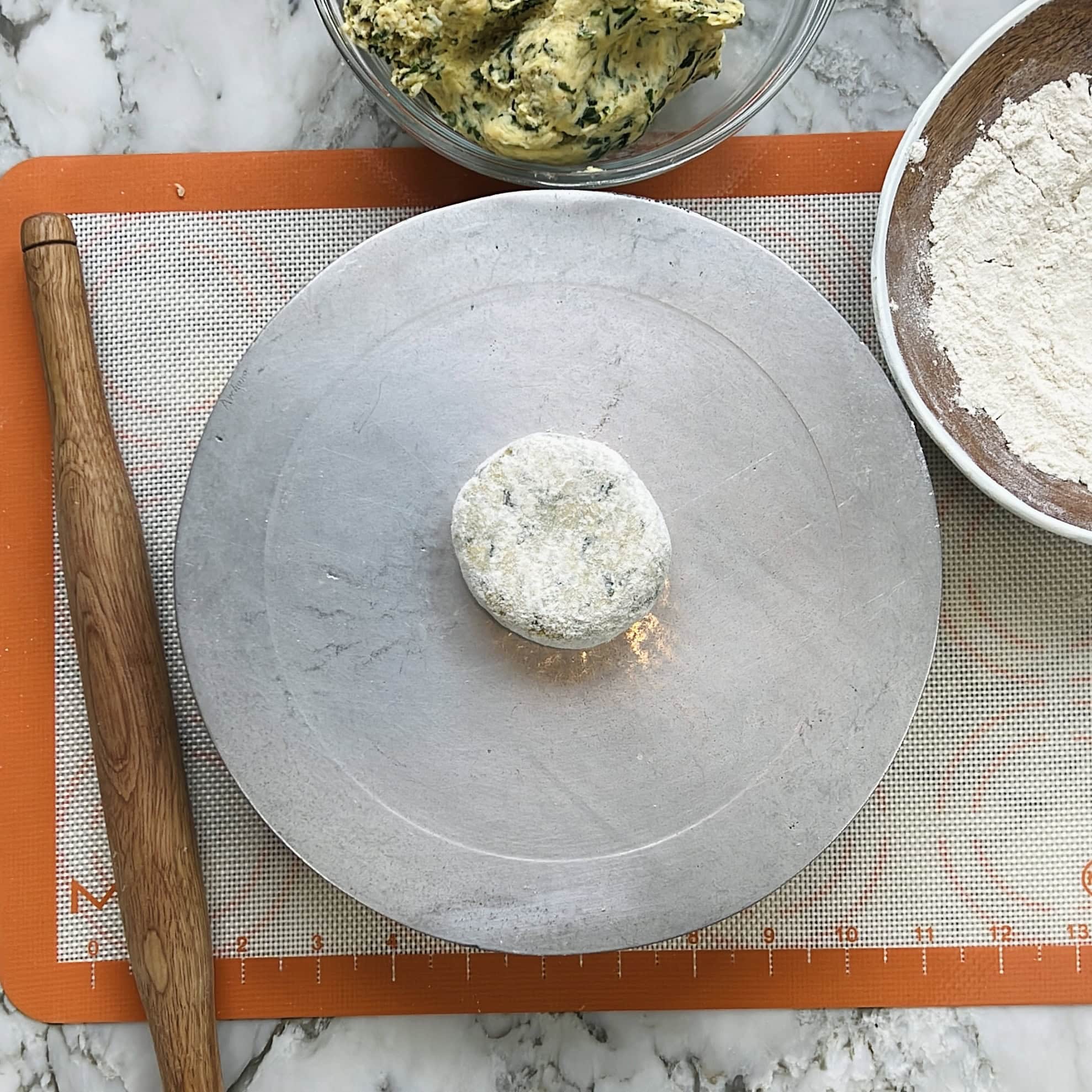 A ball of dough sits on a metal plate, surrounded by a rolling pin, a bowl of flour, and a bowl with more dough, on an orange silicone mat.