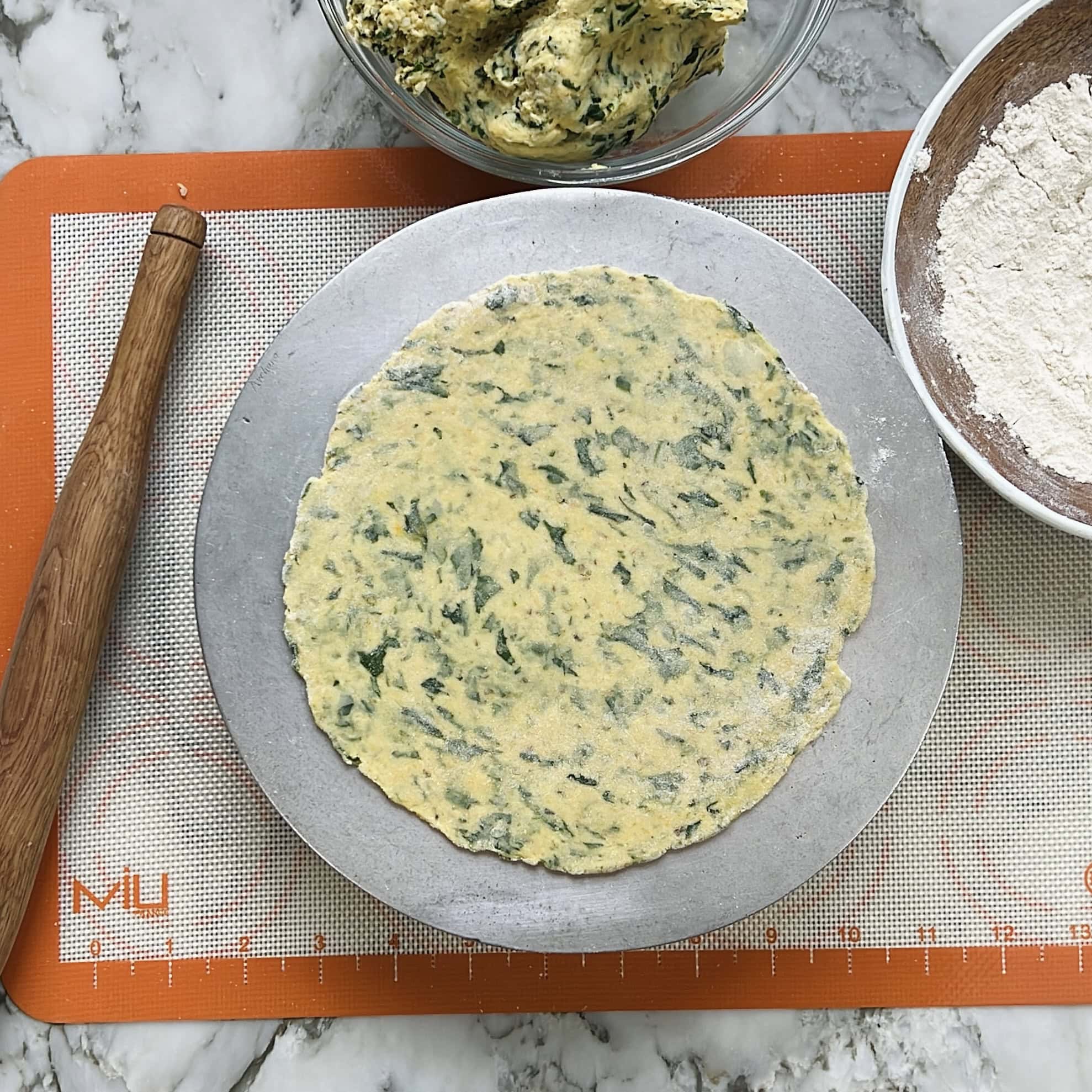 A rolled-out piece of green-flecked dough on a round metal tray, next to a wooden rolling pin, a bowl of flour, and a bowl of dough on a silicone baking mat.
