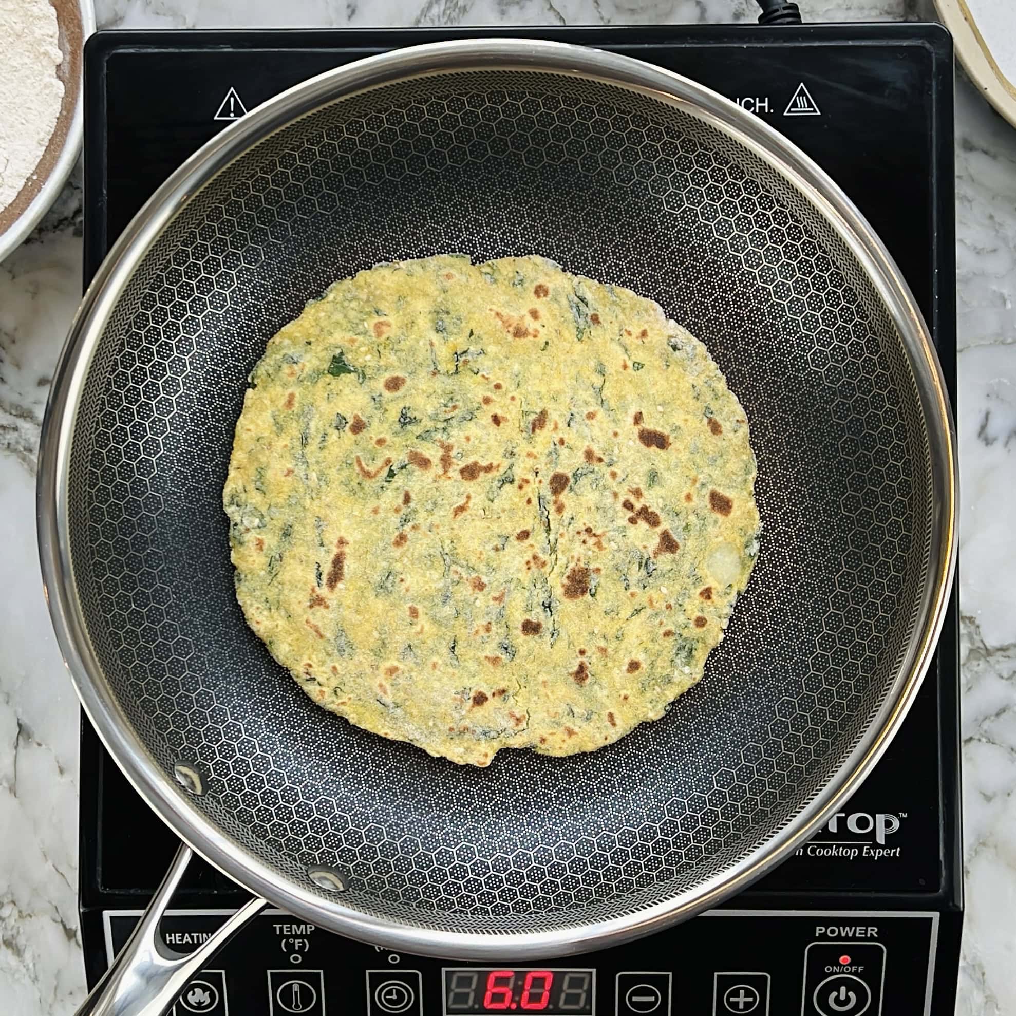 A flatbread with visible kale cooks in a nonstick pan placed on an induction cooktop.