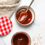 Top-down view of a jar and bowl of tamarind date chutney with a spoon, a gingham lid, and a cloth on a white surface. T