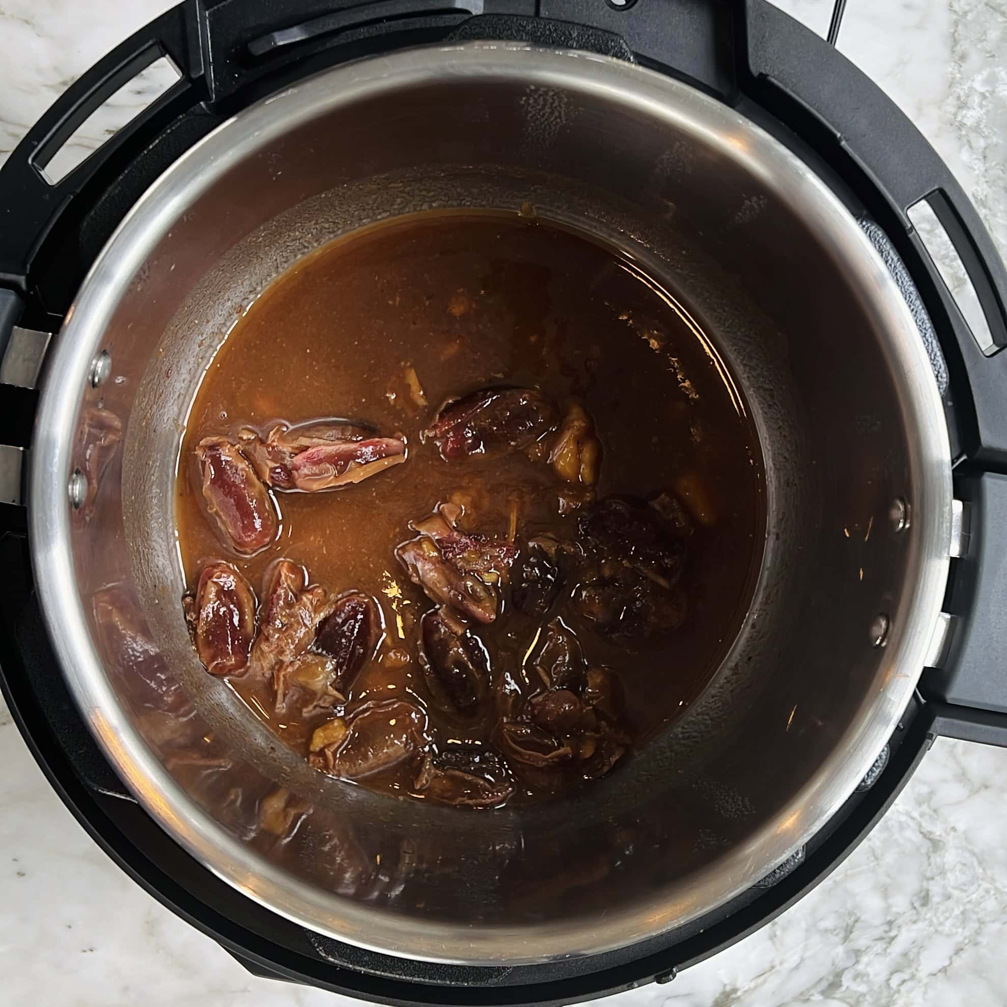 A stainless steel pot contains chopped dates simmering in a brown liquid mixture. The pot is set inside an electric pressure cooker on a marble countertop.