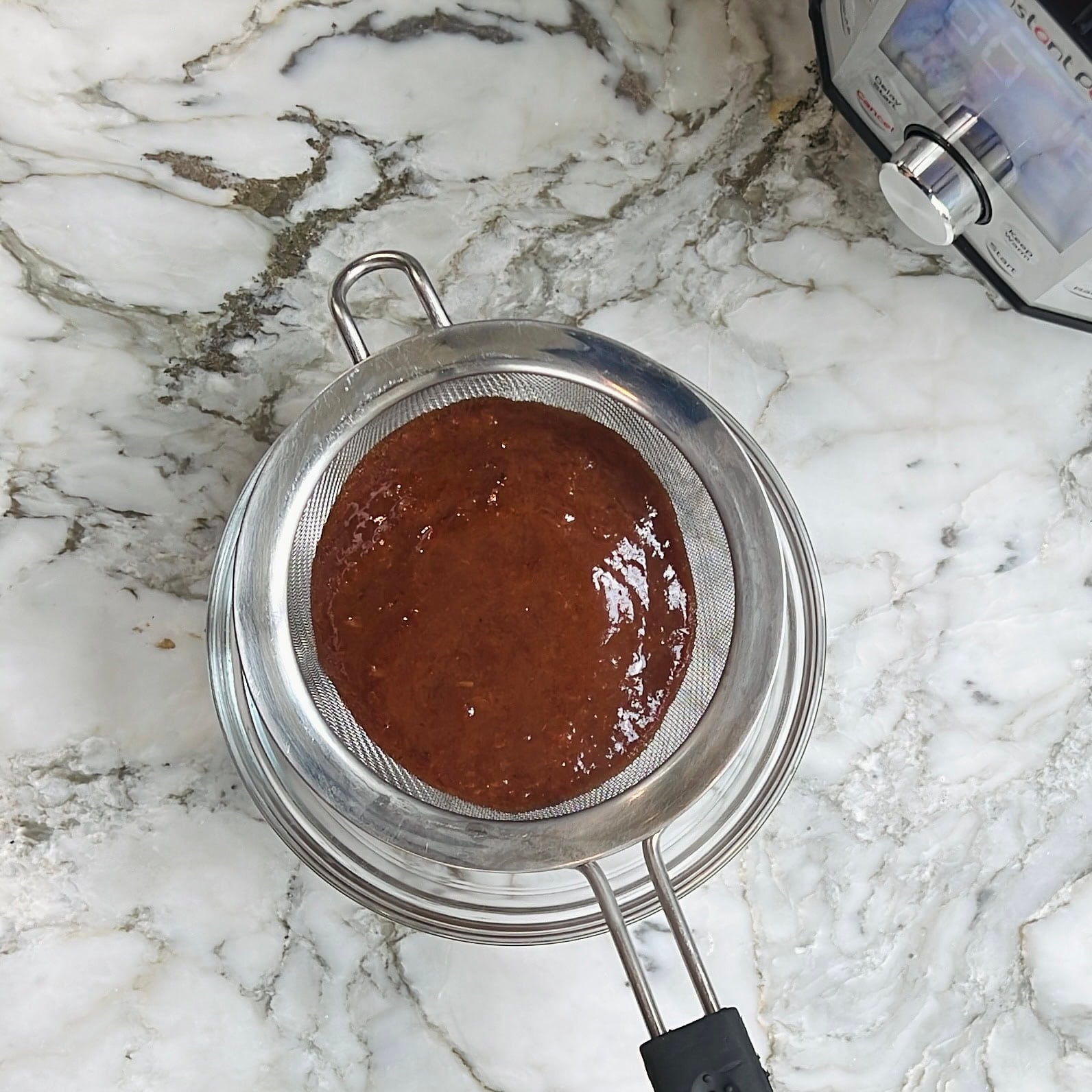 A metal strainer with thick brown sauce sits over a metal bowl on a marble countertop, next to an electric pressure cooker.