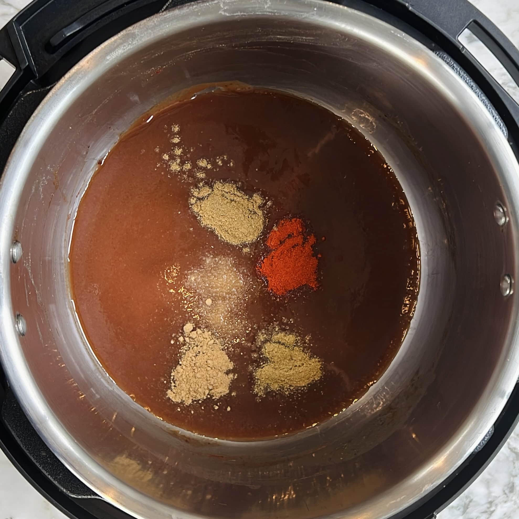 Spices and sauce are combined in an Instant Pot, with ground spices sprinkled on top of a brown liquid base.