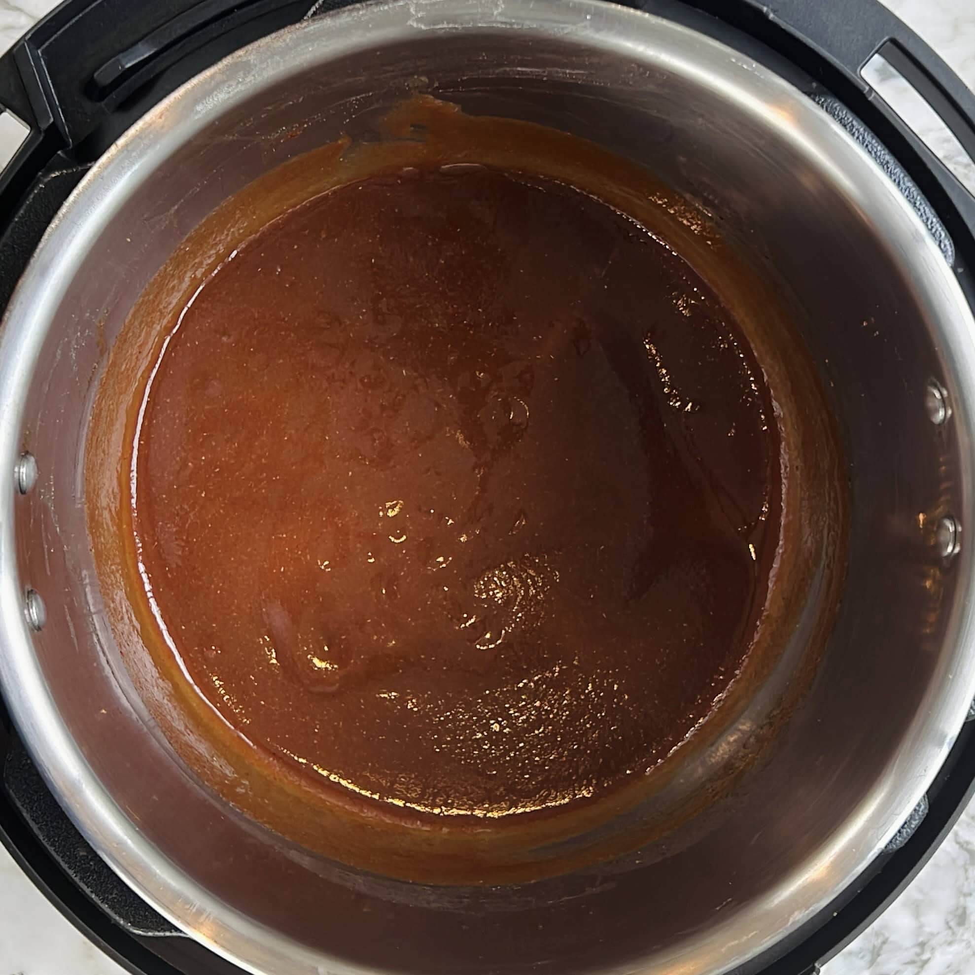 A thick, reddish-brown sauce is shown inside a stainless steel inner pot, likely part of an electric pressure cooker on a white countertop.