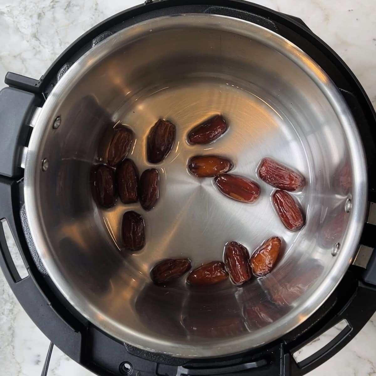 A stainless steel pot with several pitted dates and a small amount of water at the bottom, placed on a marble countertop.