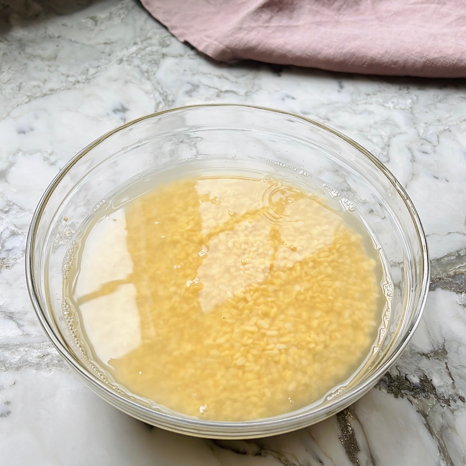 A clear glass bowl filled with lentils soaking in water sits on a marble countertop.