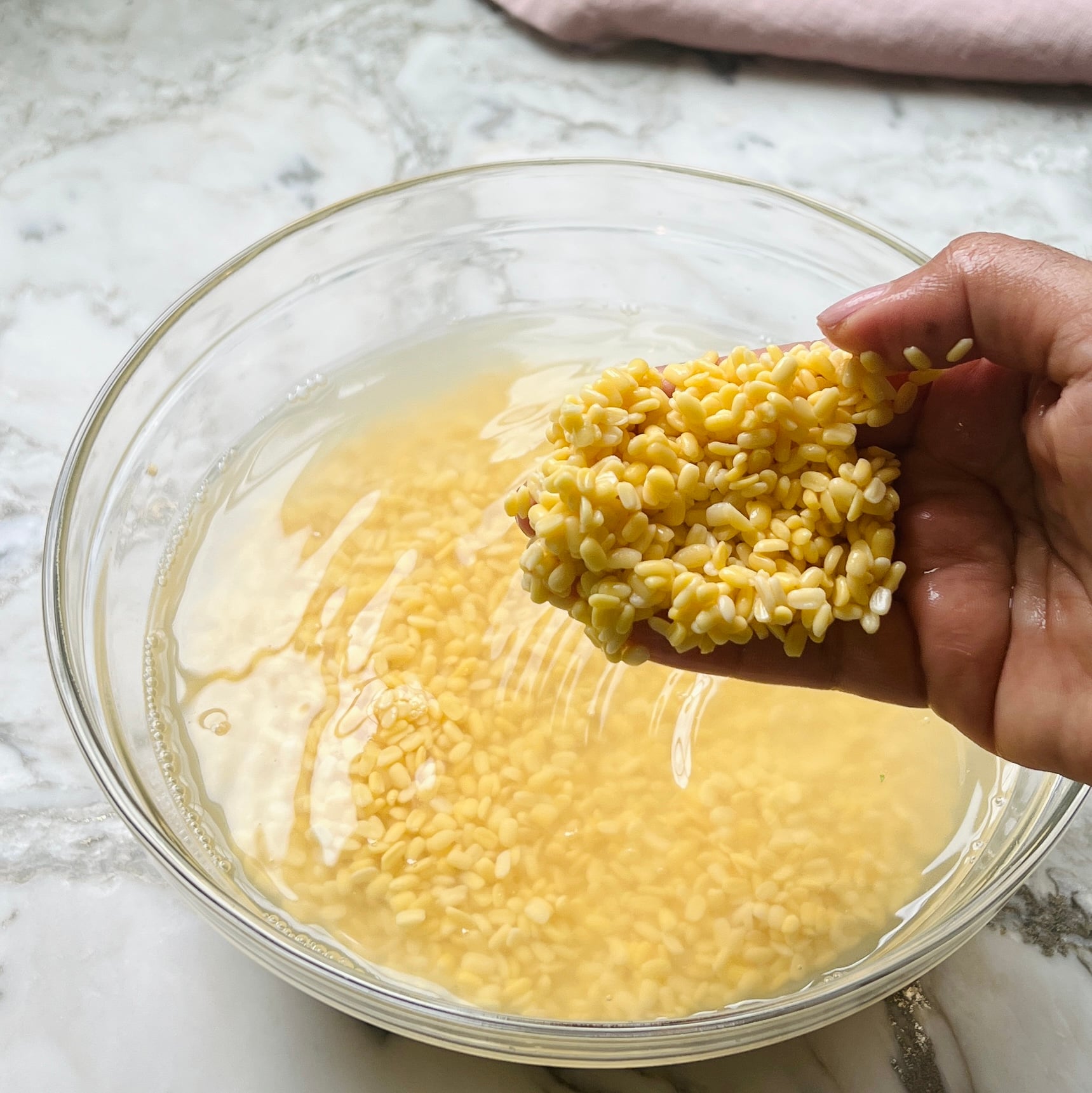 A hand holds soaked yellow lentils above a glass bowl filled with water and more lentils on a marble surface.