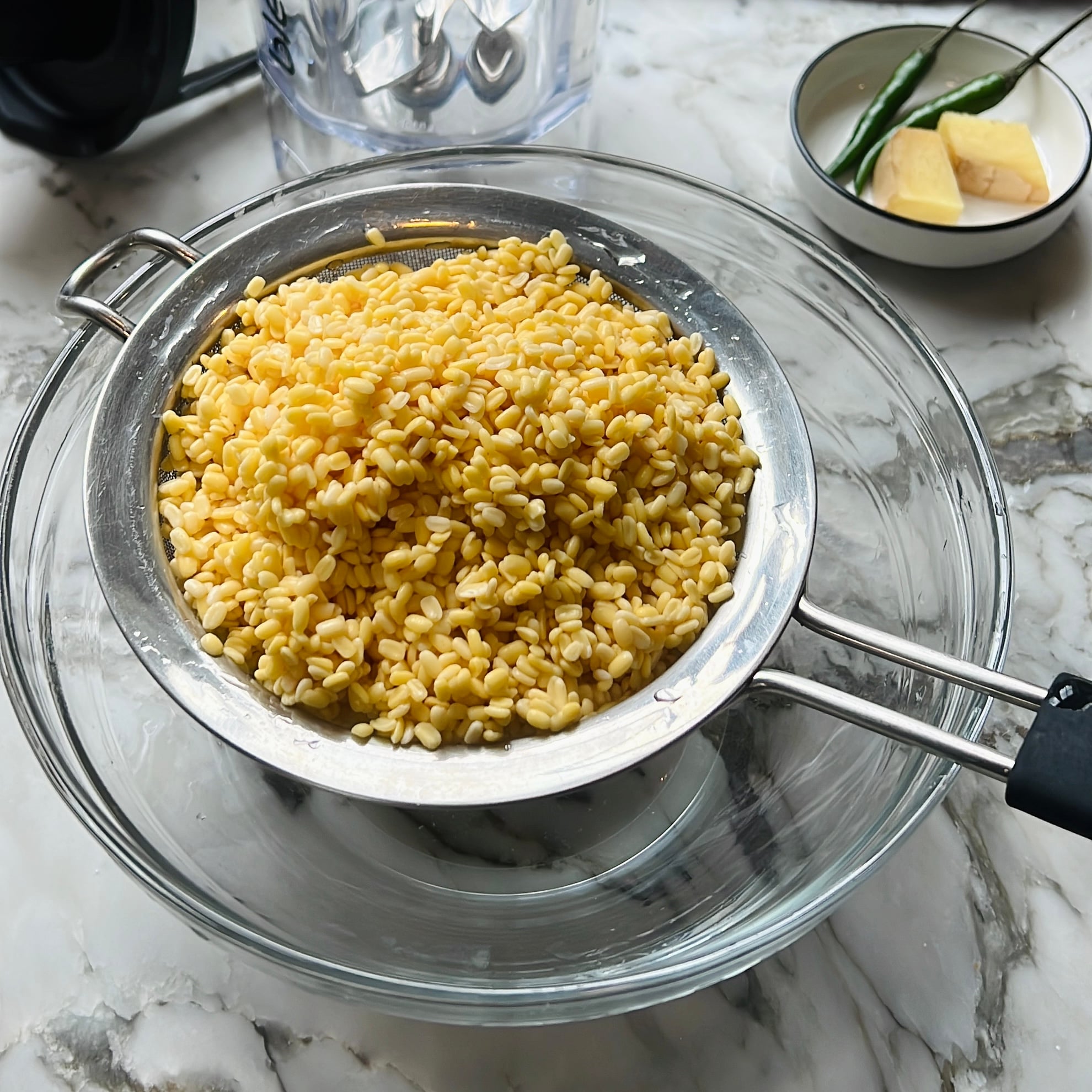 A metal strainer with soaked yellow lentils sits over a glass bowl on a marble counter.