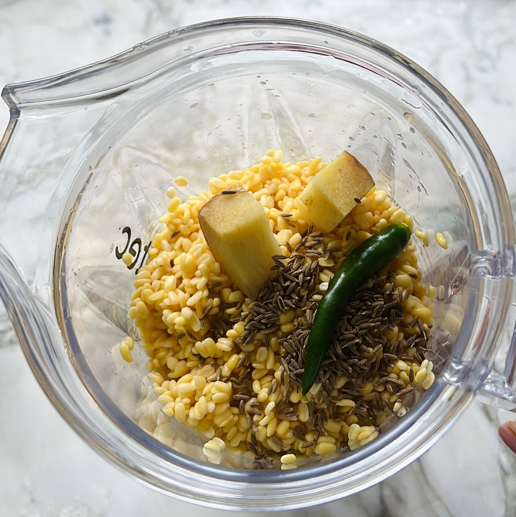 A blender contains soaked yellow lentils, two ginger pieces, a green chili, and cumin seeds, ready to be blended.