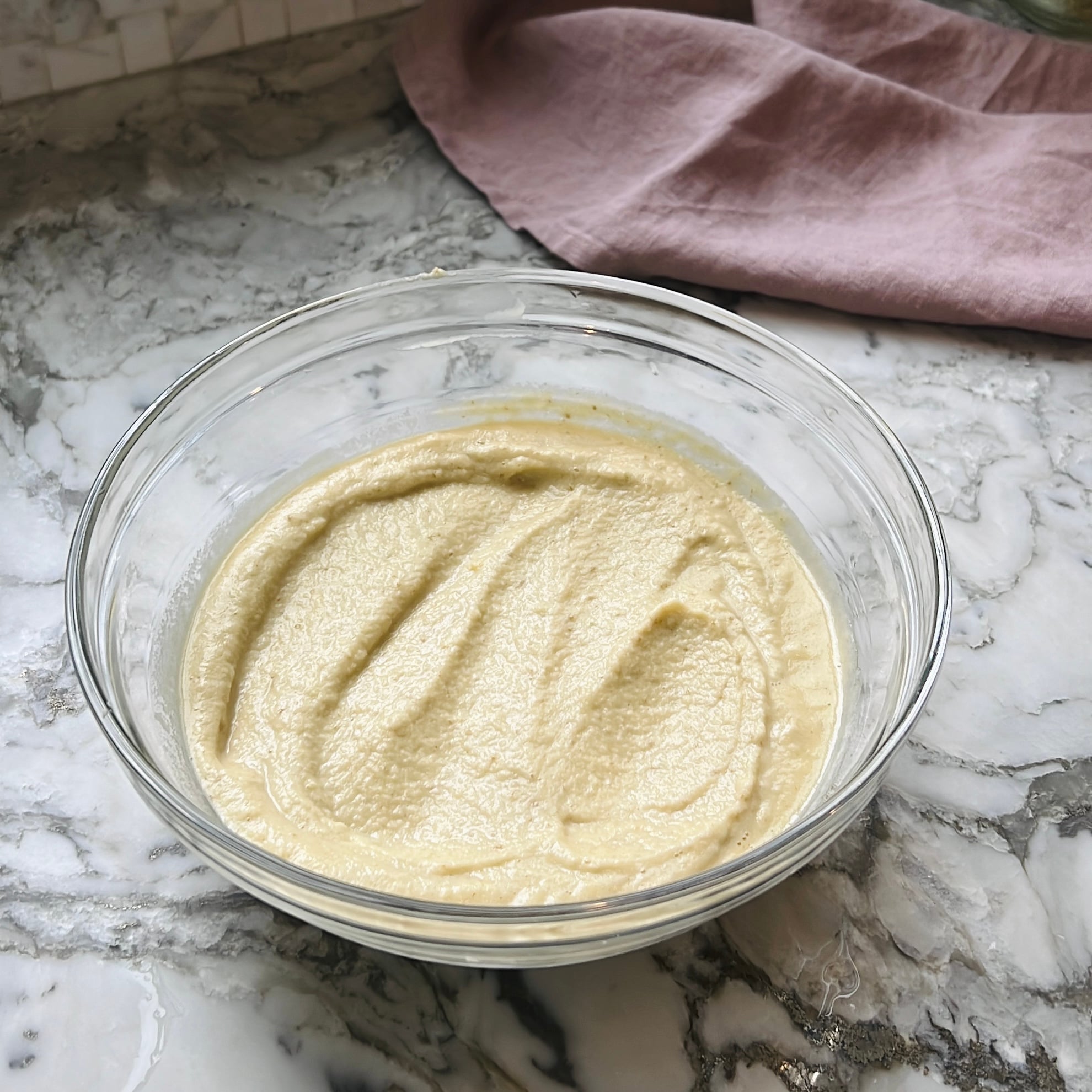 A glass bowl filled with creamy lentil batter sits on a marble countertop.