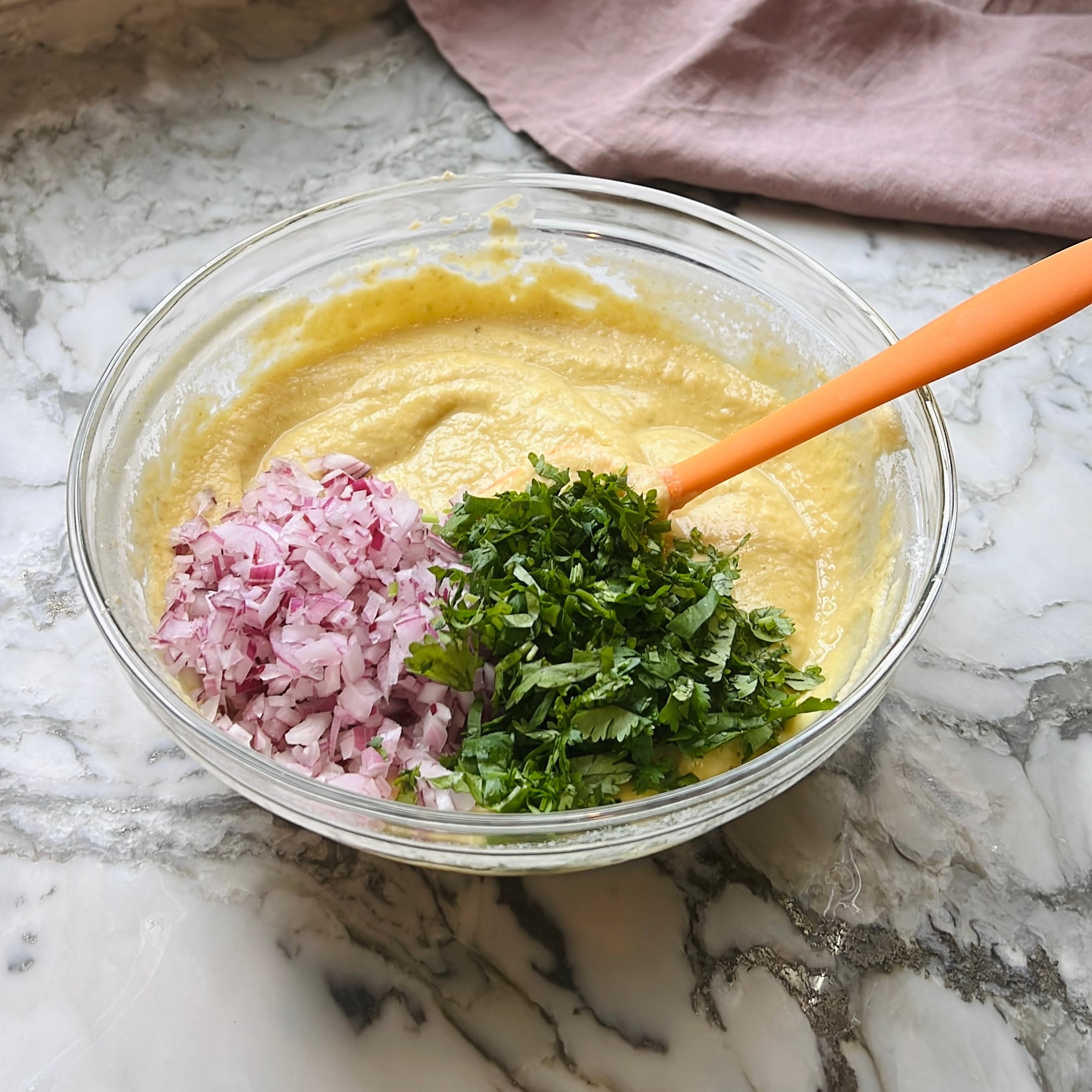 A glass bowl with mung dal batter, chopped red onions, and cilantro, with an orange spatula mixing the ingredients.