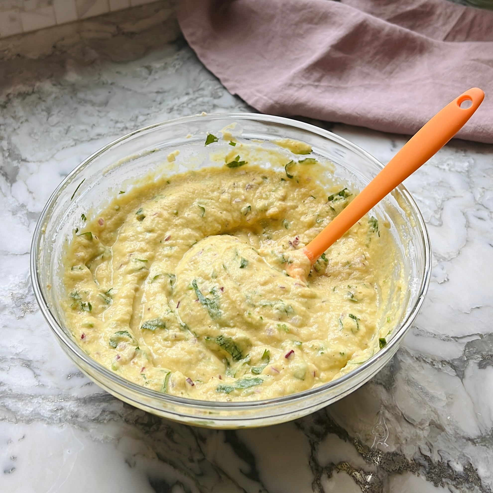 A glass bowl of mixed batter with visible herbs and spices sits on a marble counter, with an orange spatula resting in the bowl.