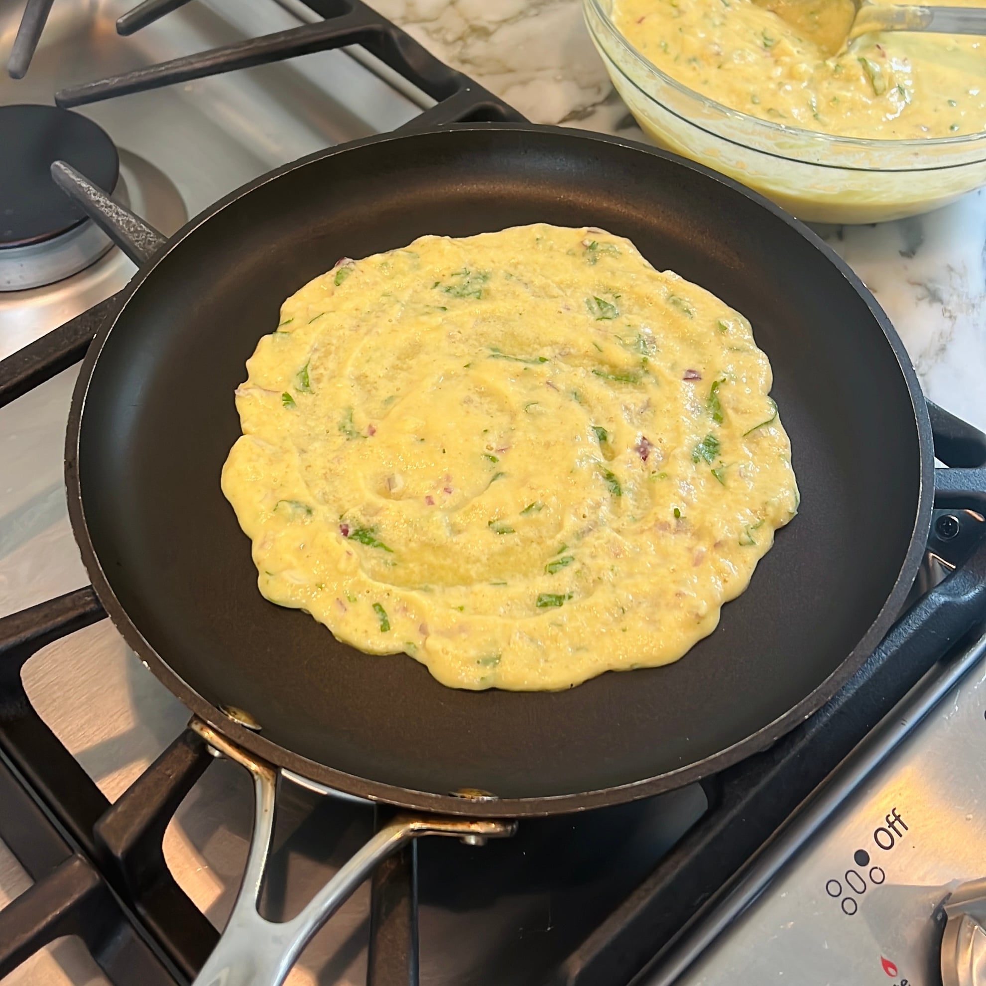 Uncooked moong dal chilla batter with visible herbs and vegetables spreads in a nonstick pan on a stovetop.