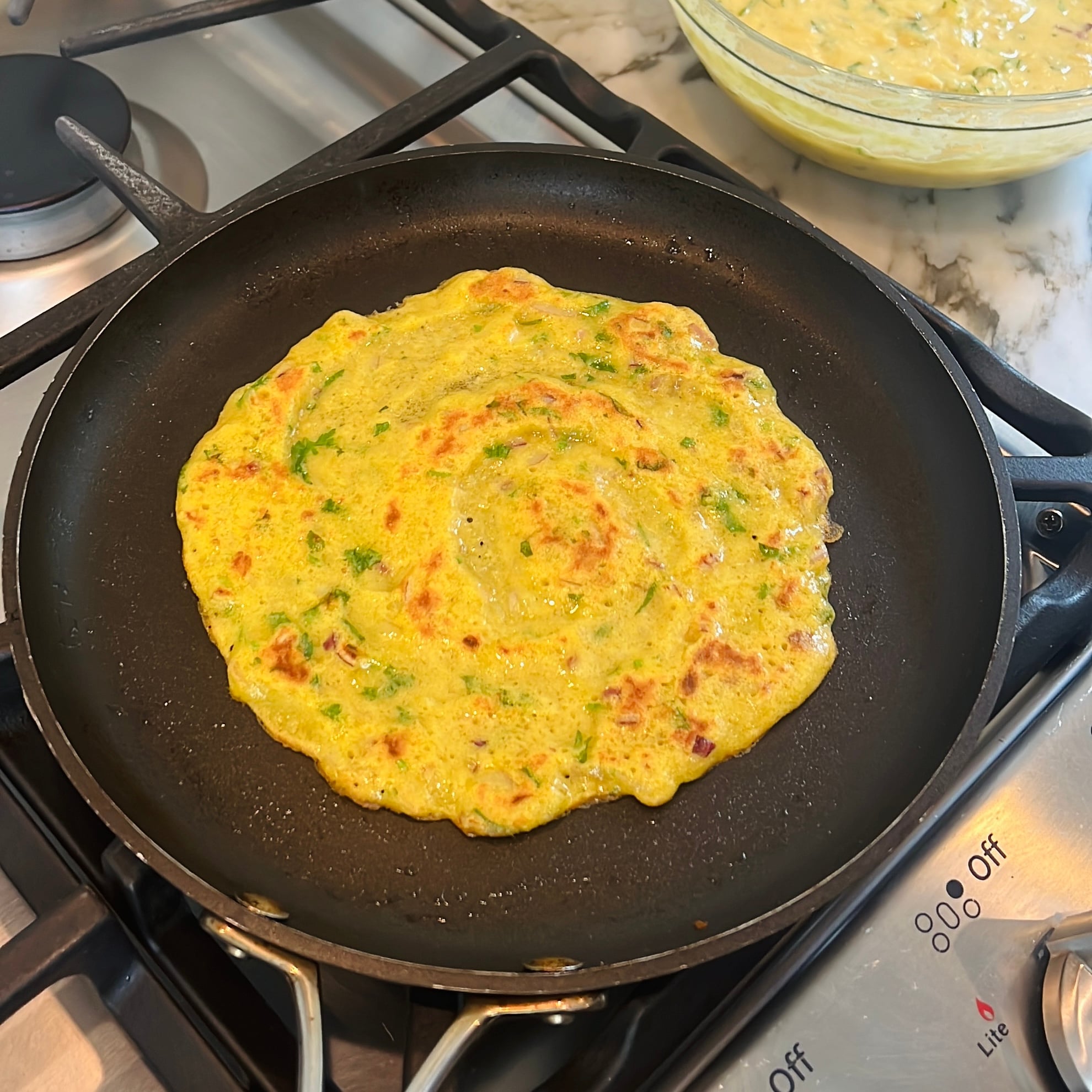 Chilla with herbs cooks in a black frying pan on a stovetop.