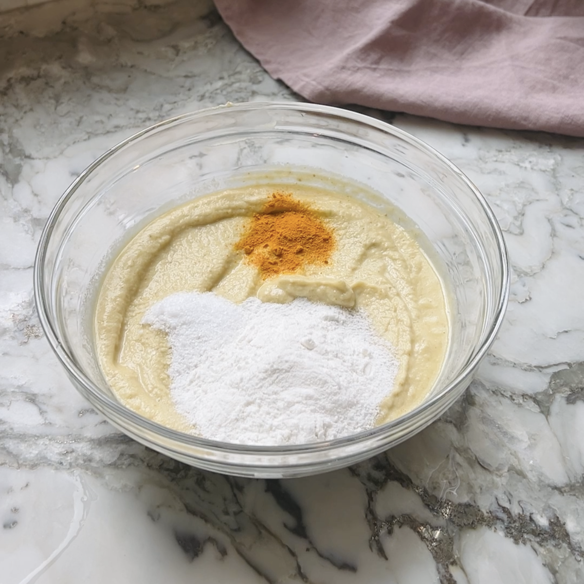 A glass bowl with batter on a marble counter, topped with rice flour and turmeric.