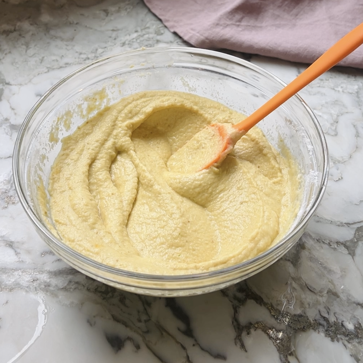 A glass bowl of yellow batter is mixed with an orange spatula on a marble countertop.
