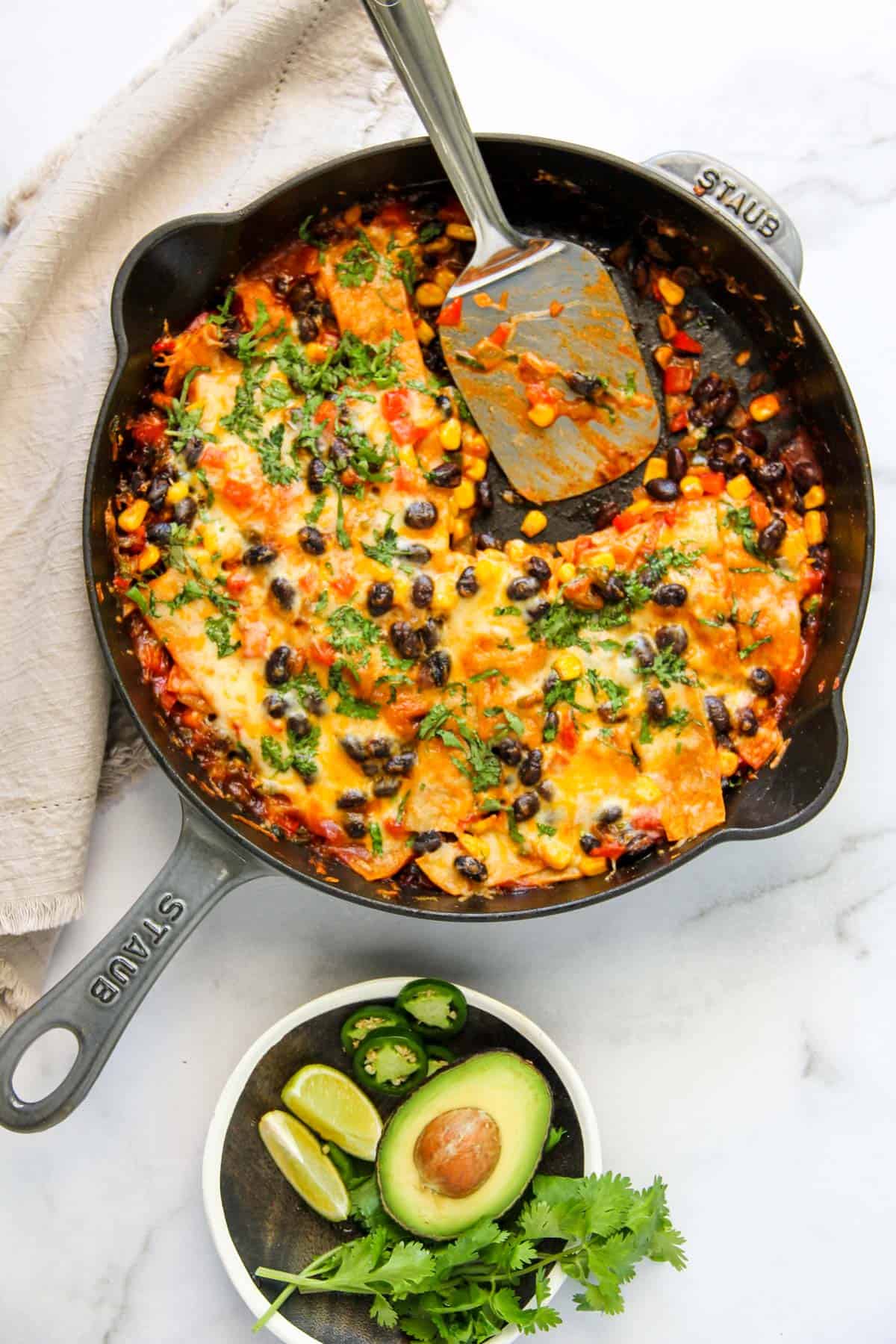 enchilada casserole in an enameled  cast iron pan with avocado, lime wedges, cilantro on the side 
