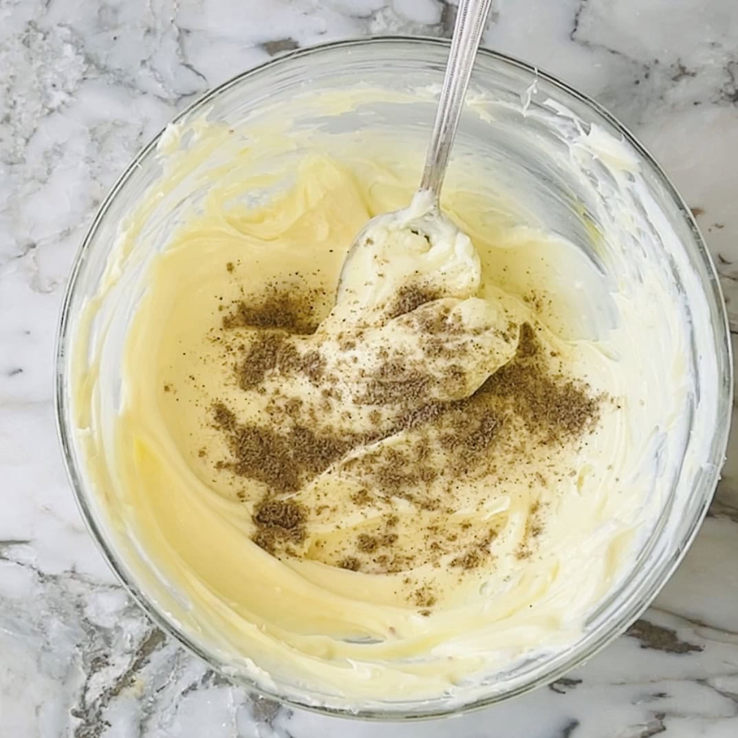 A glass bowl of creamy yellow mixture topped with ground spices sits on a marble surface, with a metal spoon resting inside.