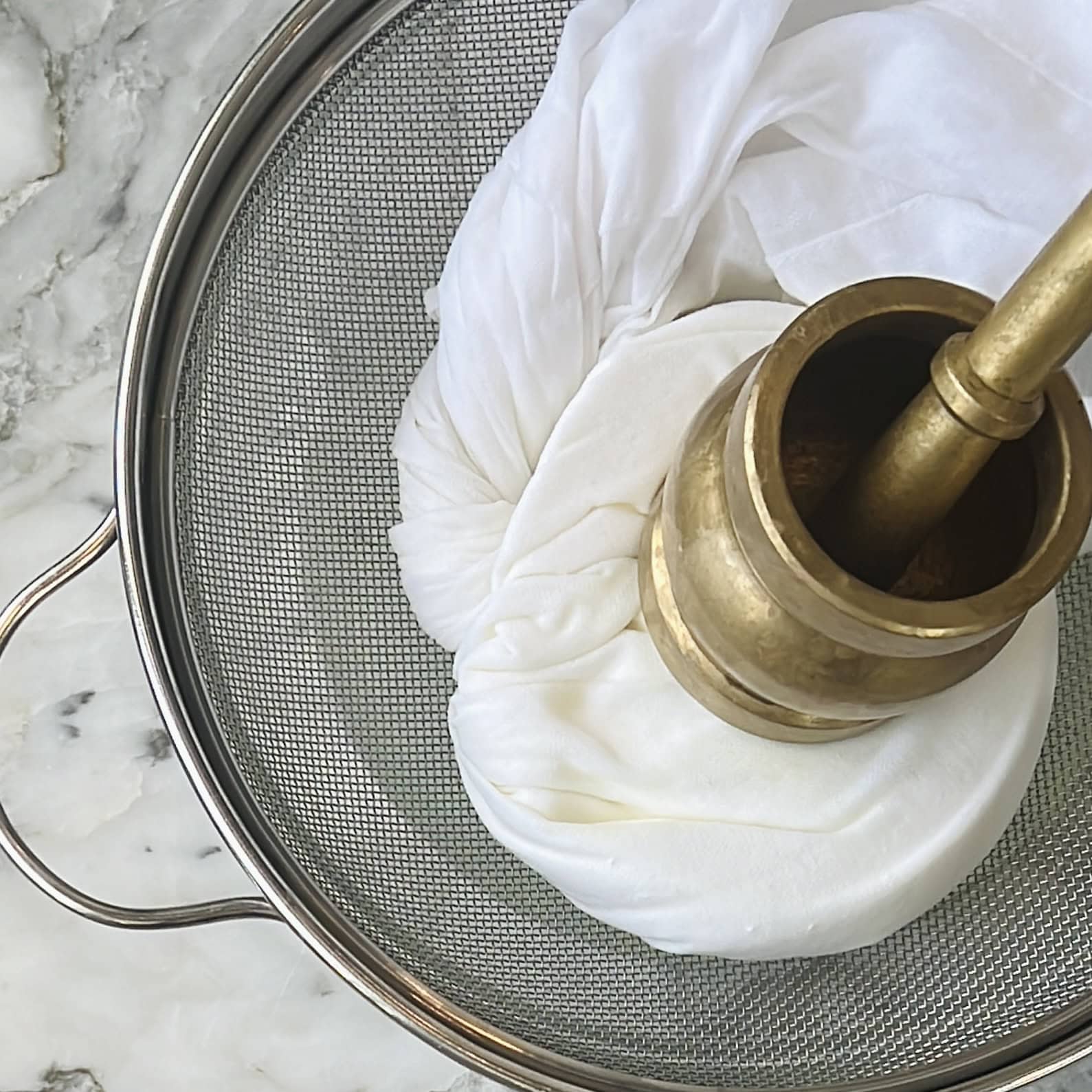A marble mortar and pestle placed on a bundle of cloth inside a metal strainer on a marble countertop.