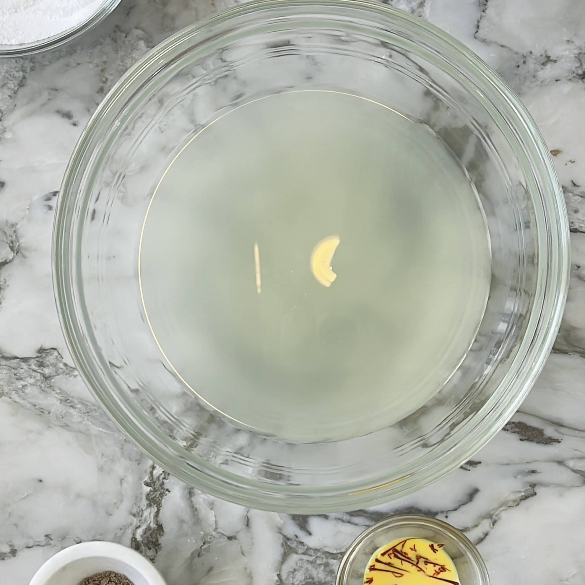 A clear glass bowl with a pale liquid and a piece of garlic, placed on a marble countertop with small bowls of spices and salt nearby.