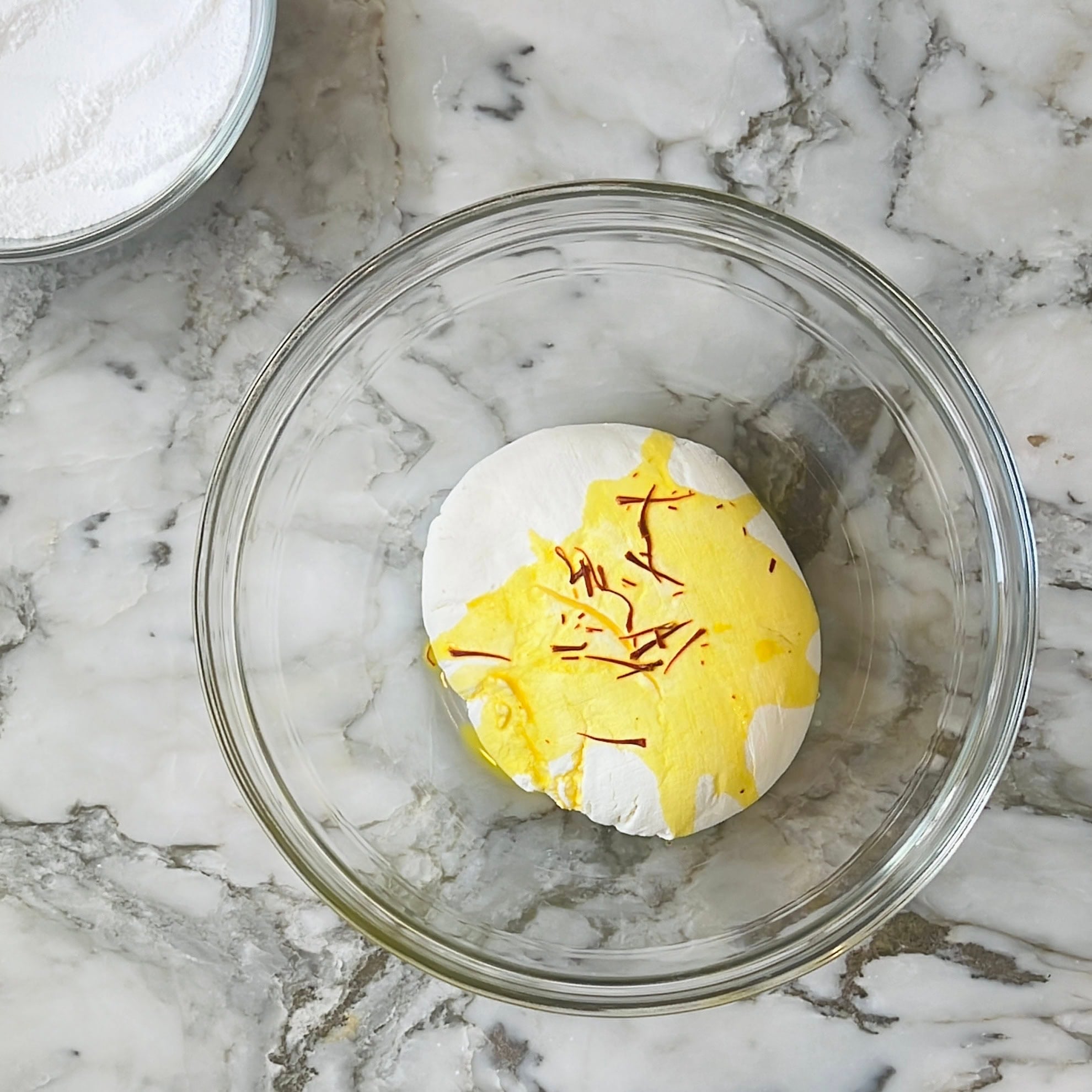 A clear glass bowl on a marble countertop contains white dough topped with yellow liquid and saffron strands. Another bowl with white powder is partially visible in the corner.
