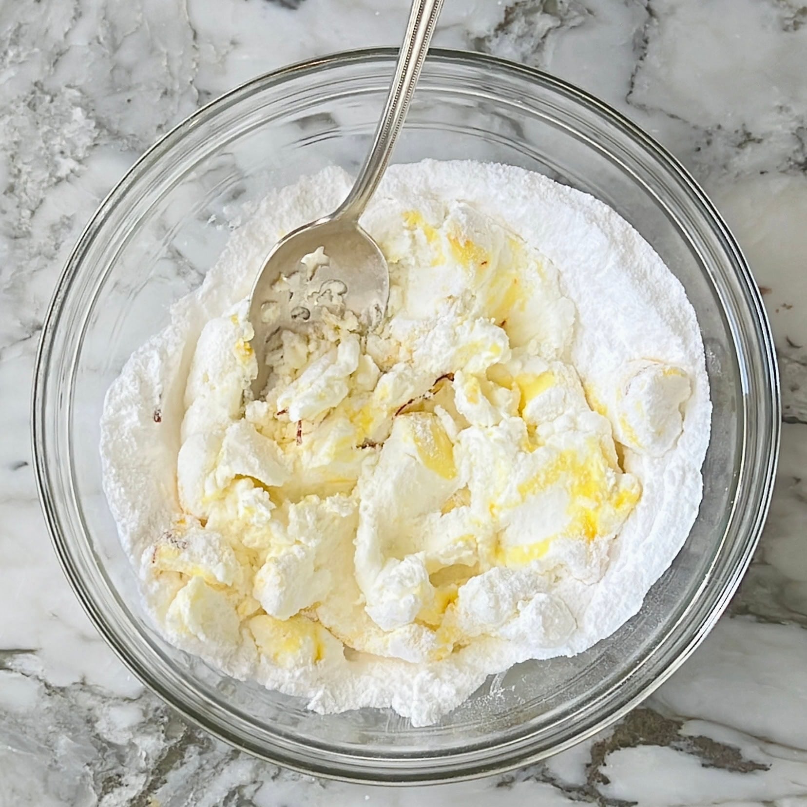 A glass bowl on a marble surface contains powdered sugar, butter, and cream cheese being mixed with a metal spoon.