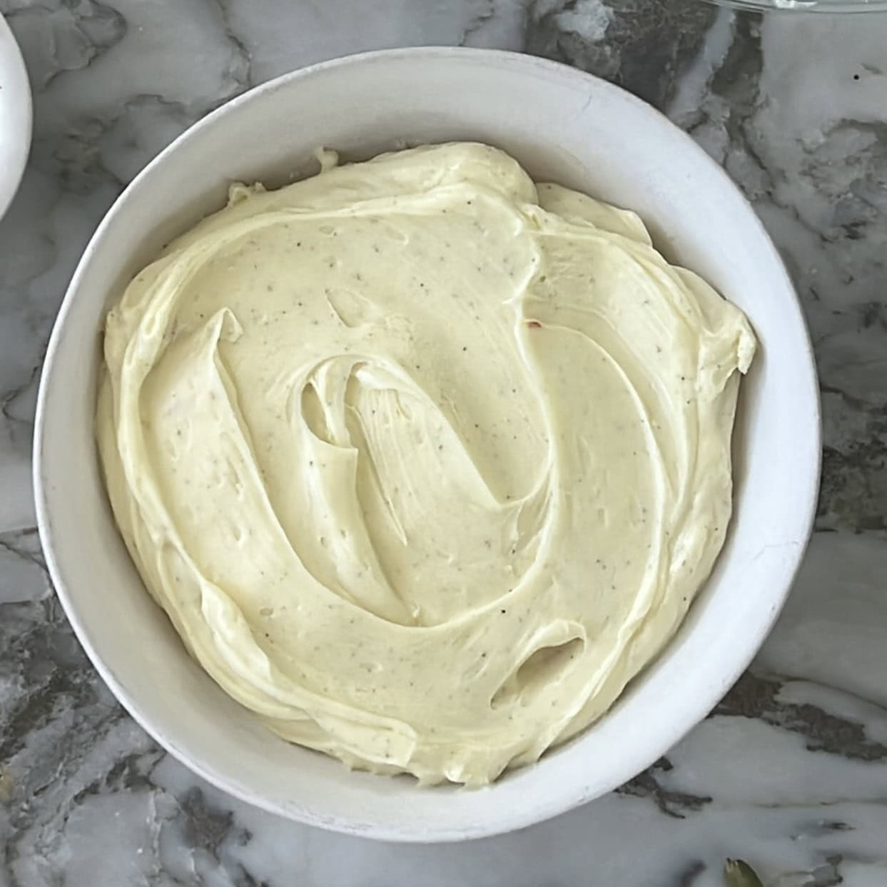 A white bowl filled with smooth, pale yellow cream cheese frosting sits on a gray marble surface.