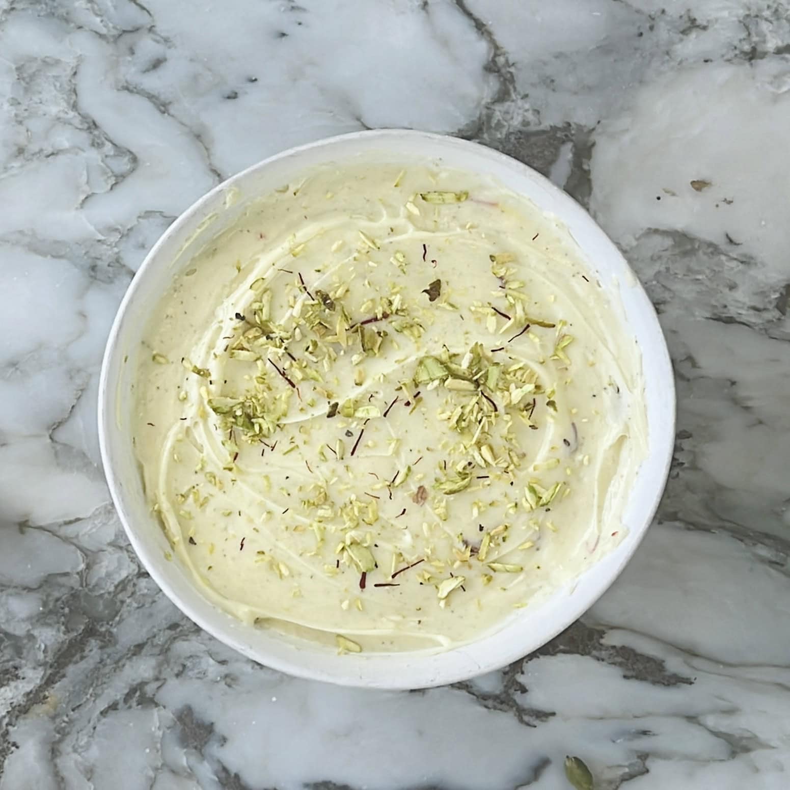 A bowl of creamy dessert garnished with chopped pistachios and strands of saffron, placed on a marble surface.