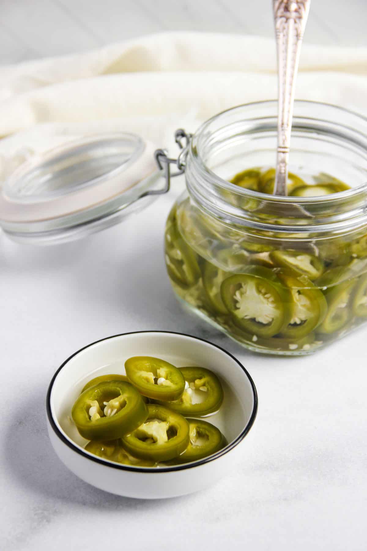 pickled jalapeños in a glass jar and a small white dish 