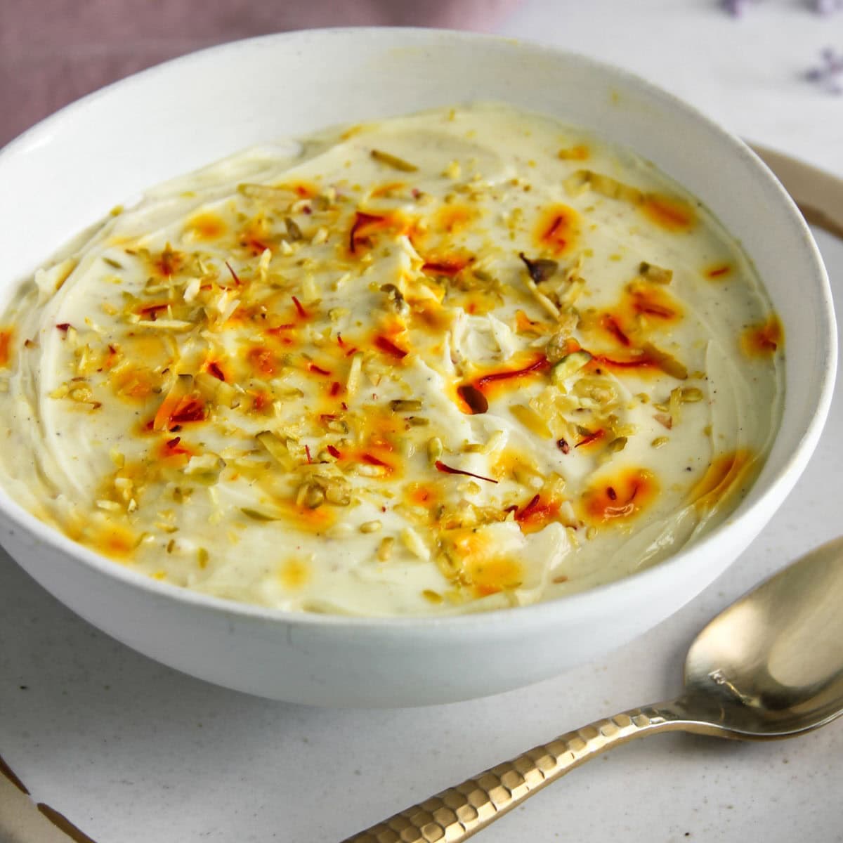 A white bowl of creamy yogurt garnished with saffron strands, chopped nuts, and a drizzle of syrup, with a spoon placed beside it.