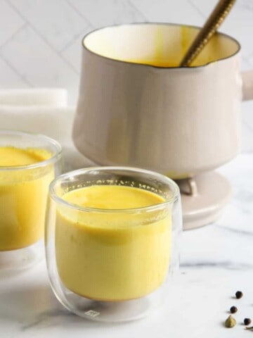 haldi doodh in two glasses with a pot in the background