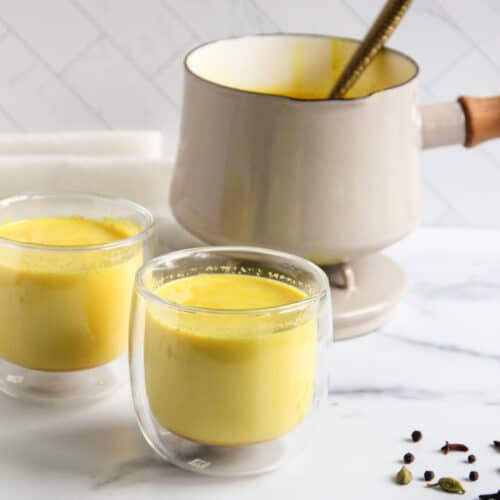 haldi doodh in two glasses with a pot in the background