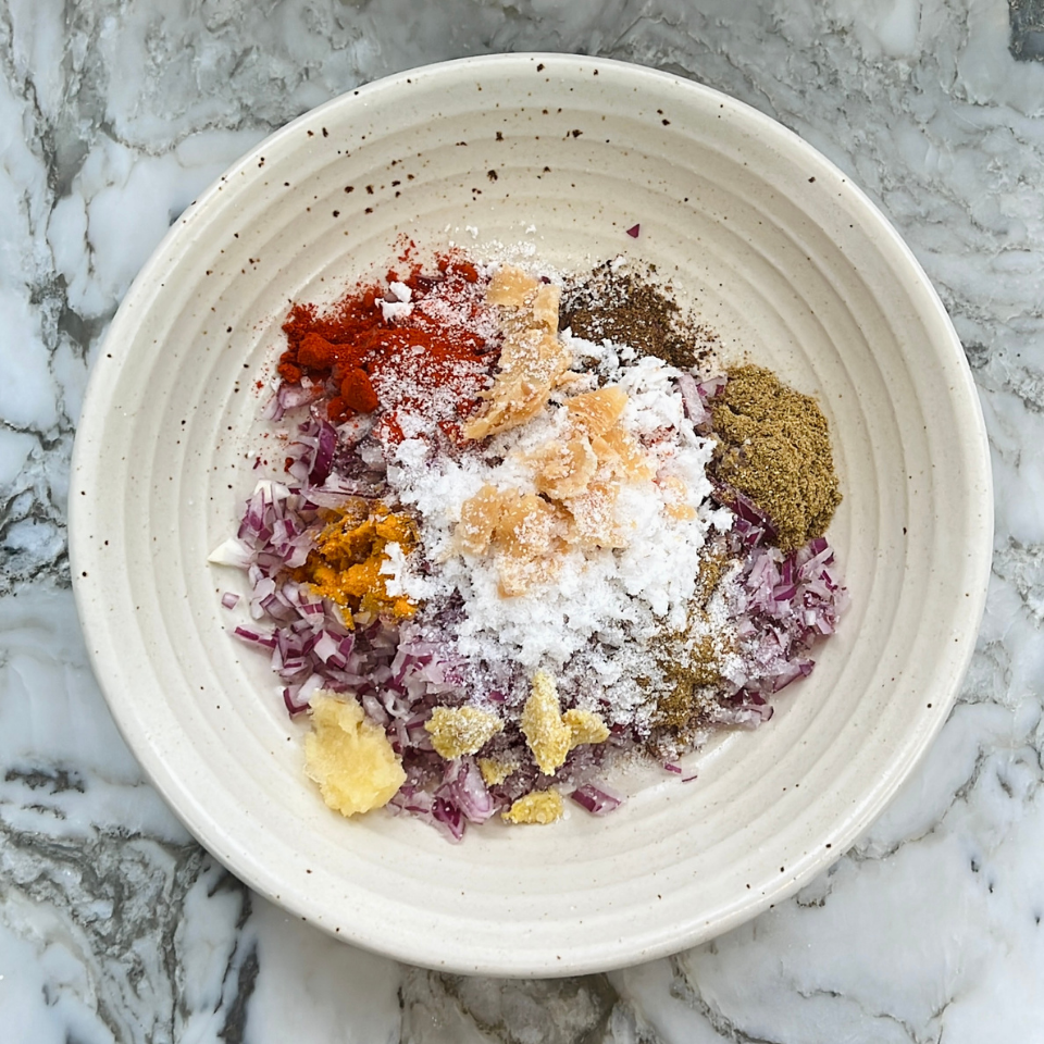 A bowl with chopped red onions, ground spices, grated ginger, turmeric, and salt on a marble surface.