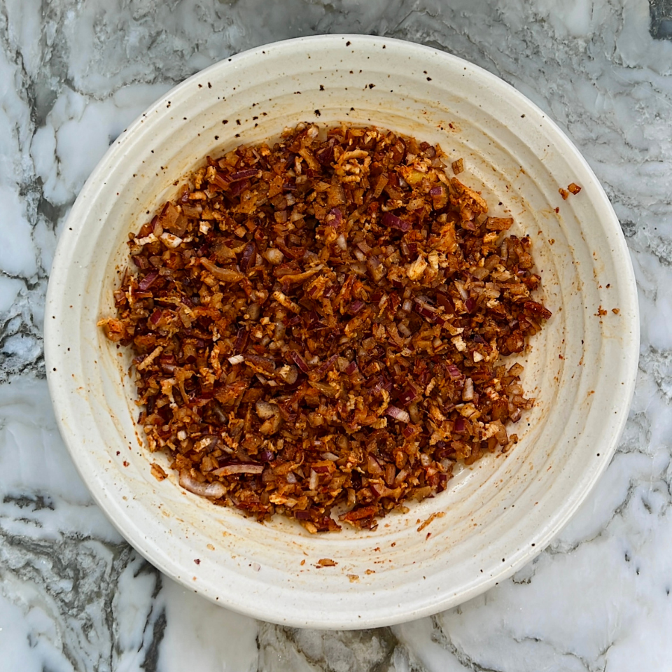 A ceramic bowl filled with a chopped mixture of onions and red spices sits on a gray and white marble surface.