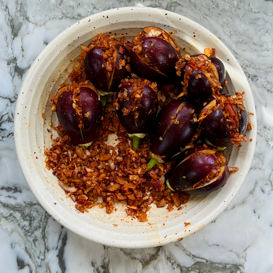 A white bowl with small eggplants stuffed with a spiced onion mixture, placed on a marble surface.