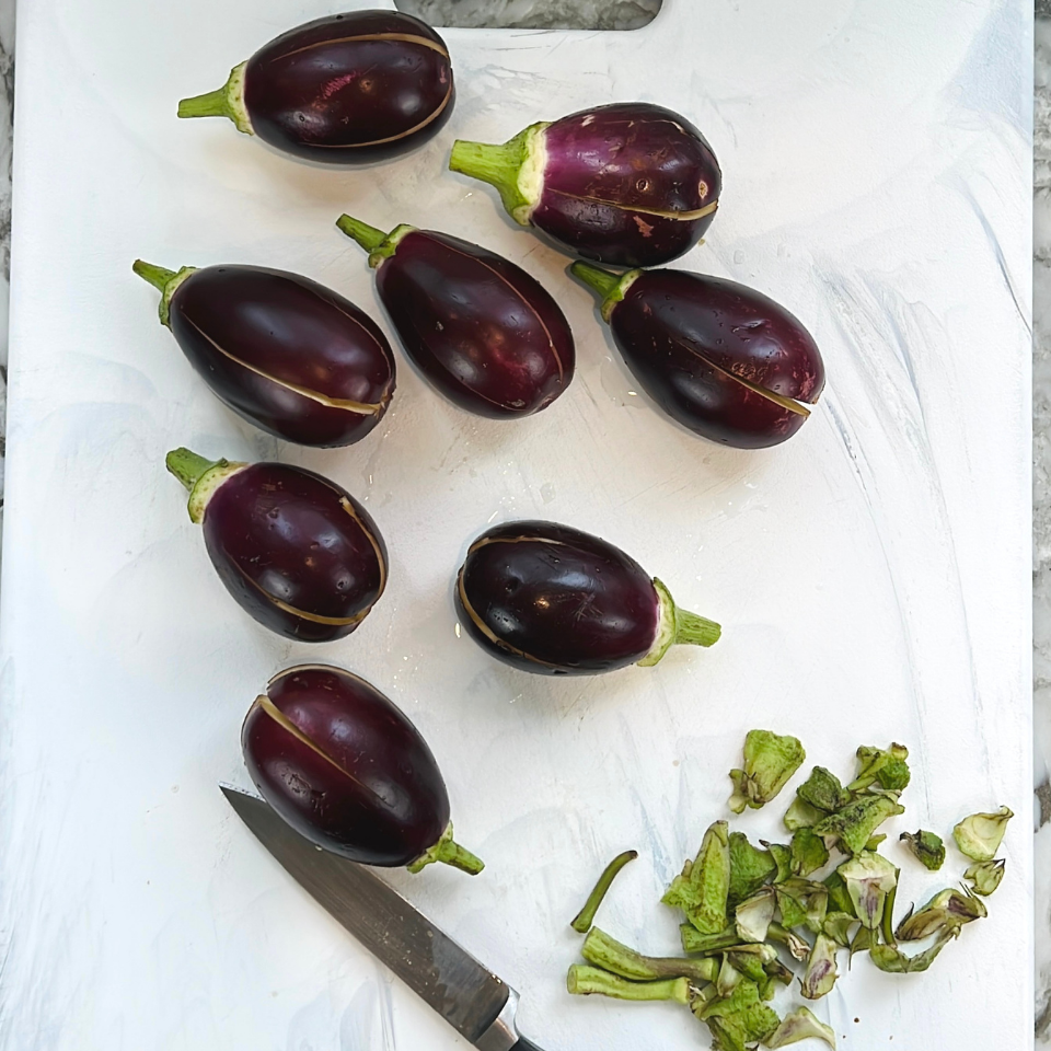 Seven small eggplants with stems partially removed are arranged on a white cutting board, with a knife and trimmed eggplant tops to the side.