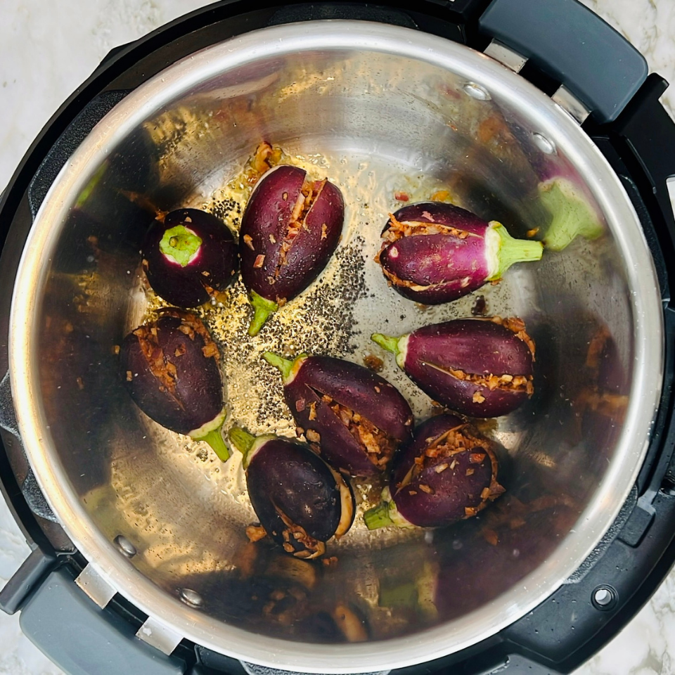 Eight whole eggplants with slits and spices are arranged inside a stainless steel pressure cooker on a countertop.