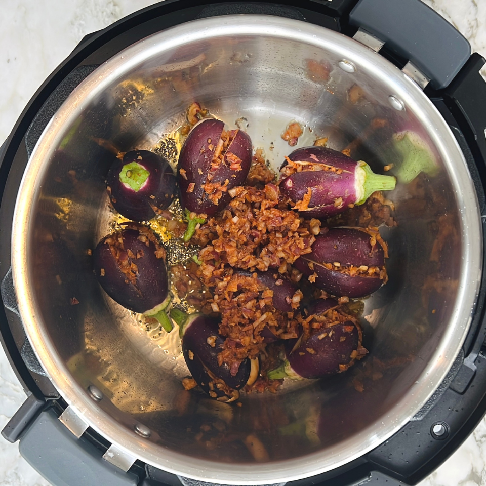 Small whole eggplants and a browned onion spice mixture in a stainless steel pressure cooker pot, ready to be cooked.