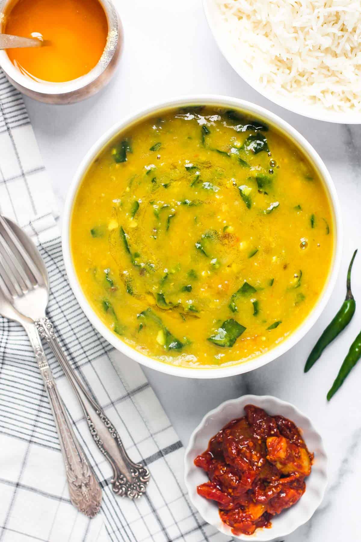 spinach dal in a white bowl along side rice, pickle and ghee bowls