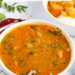 Bowl of South Indian sambar with vegetables, garnished with cilantro, a spoon inside, and a side bowl partially visible in the background.