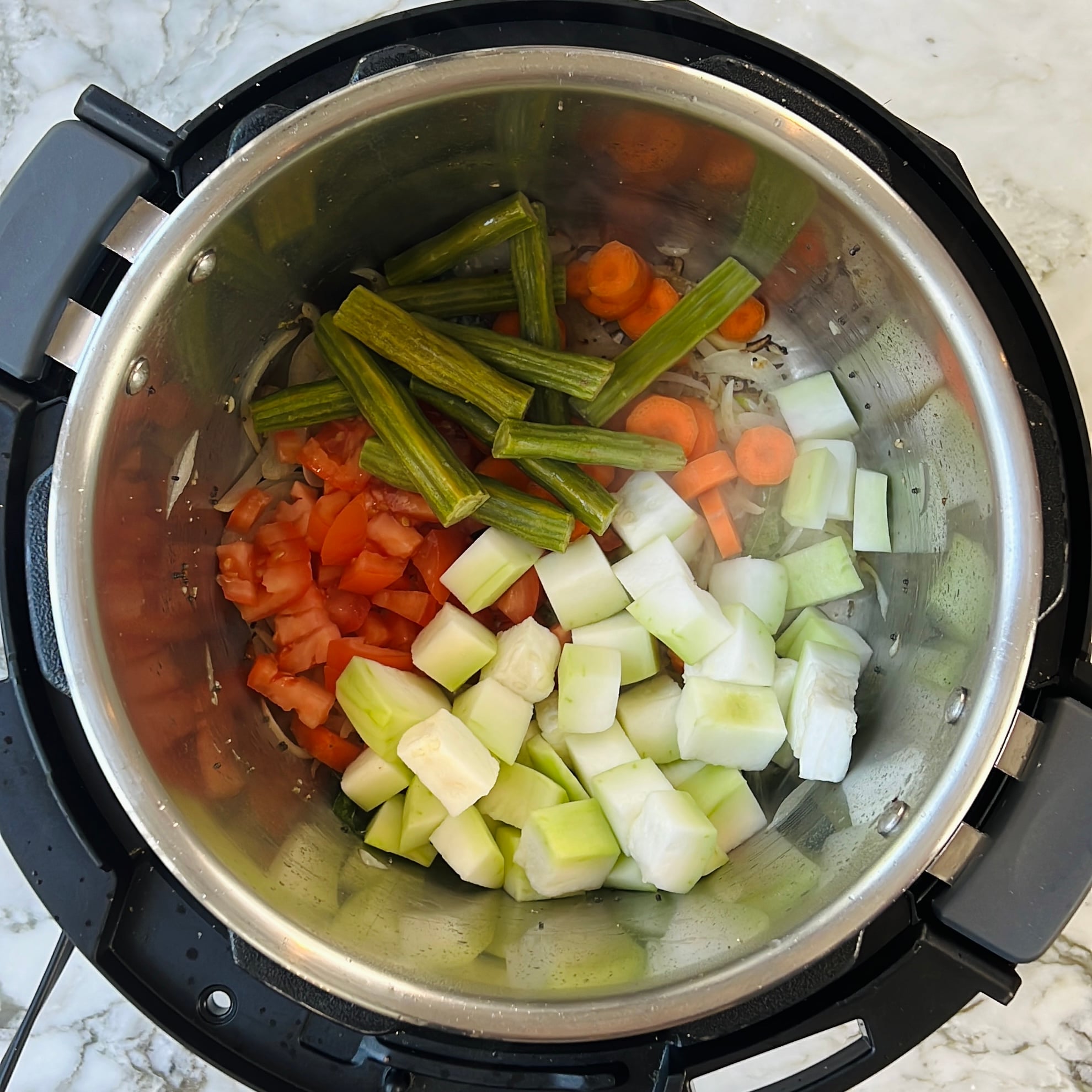 Chopped tomatoes, carrots, drumsticks, and dudhi in a pressure cooker on a marble countertop.
