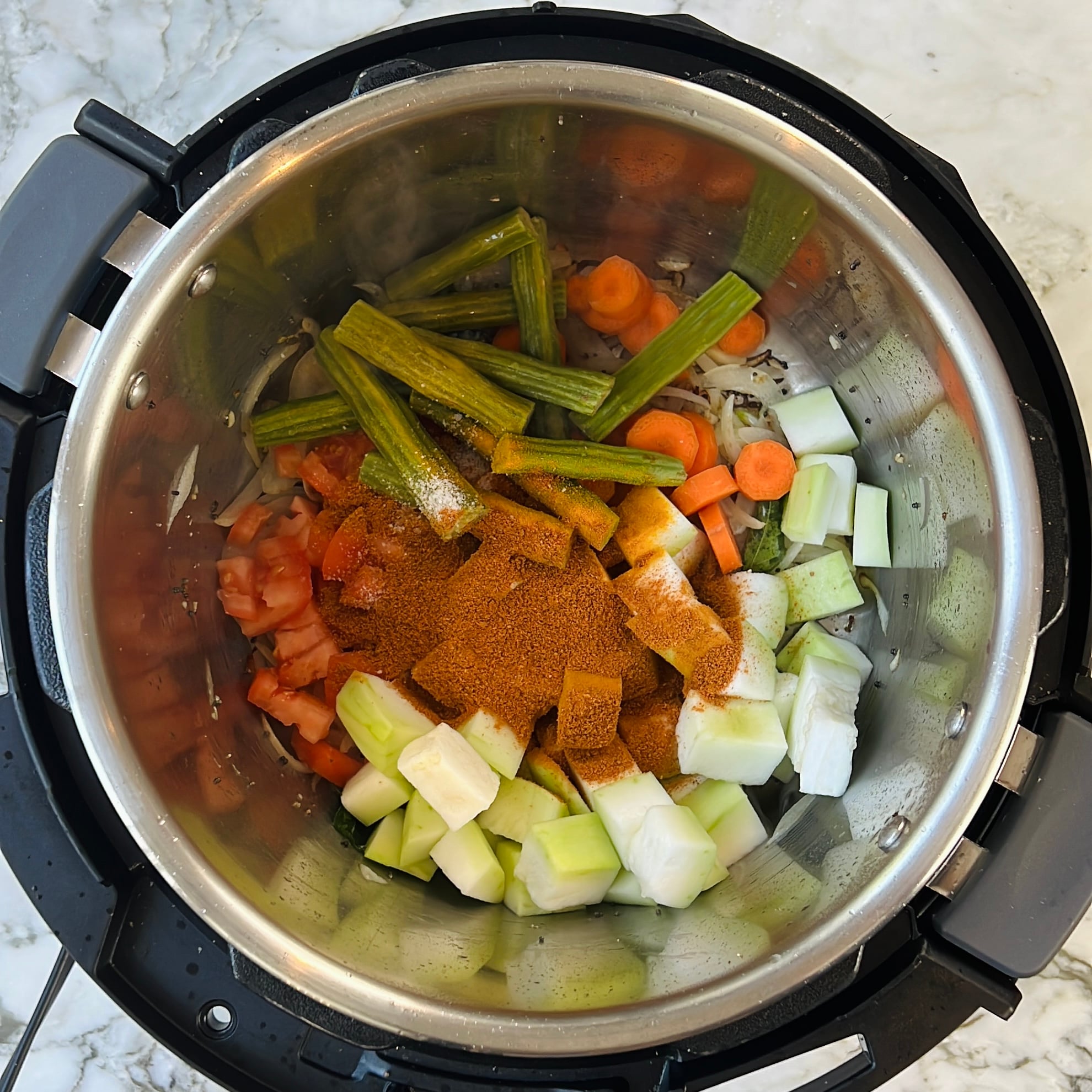 Chopped vegetables, spices, and drumsticks are placed inside an Instant Pot on a marble countertop, ready for cooking.