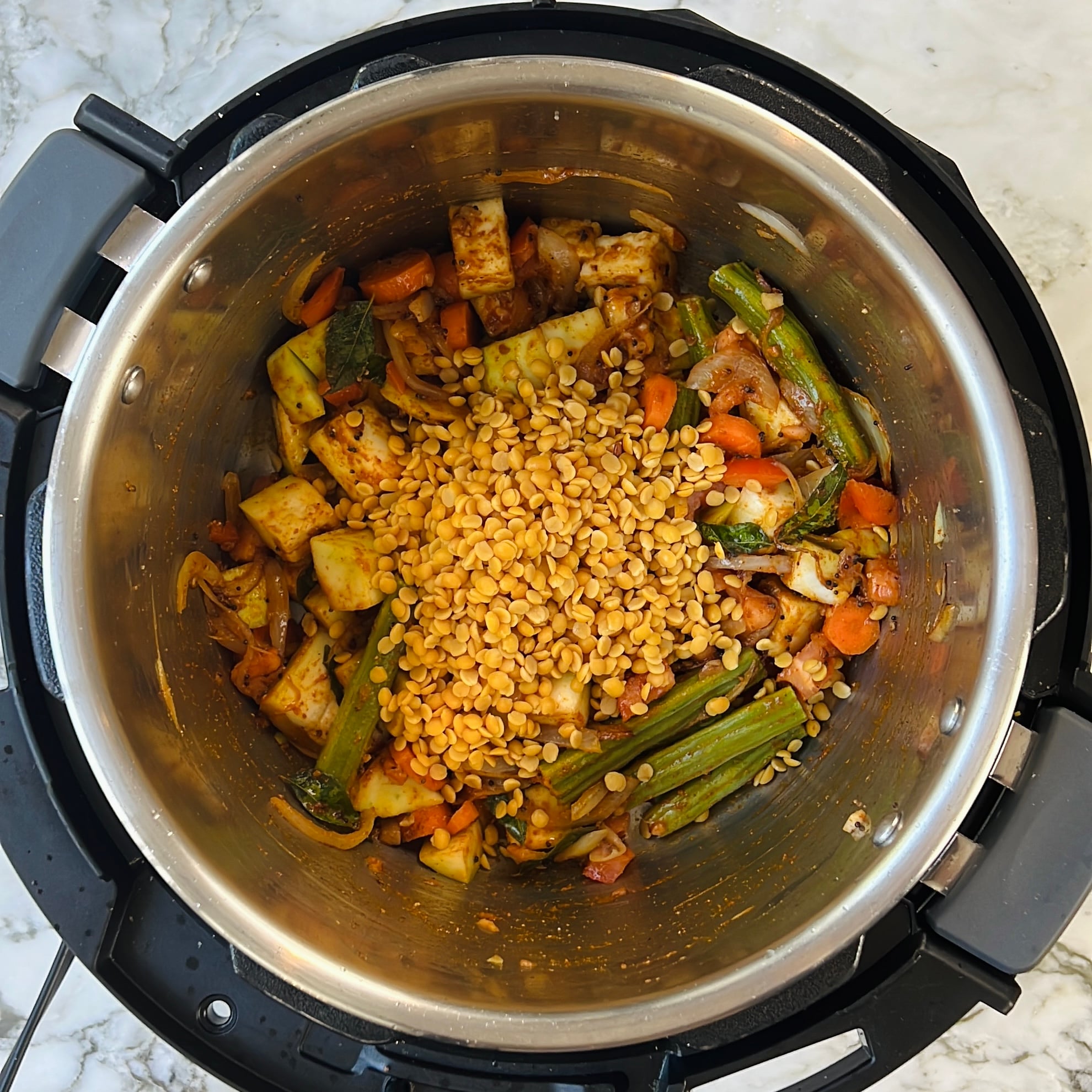 Chopped vegetables and yellow lentils in an Instant Pot, ready to be cooked, on a marble countertop.