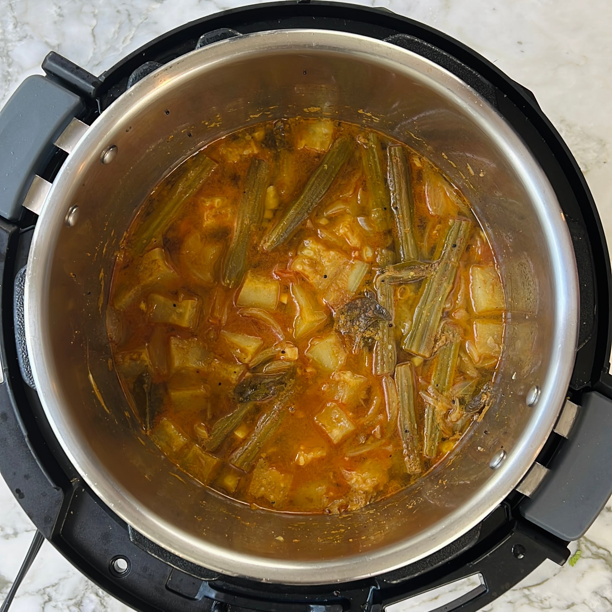 Instant pot filled with cooked vegetable sambar, including drumsticks and chunks of dudhi, sits on a marble countertop.