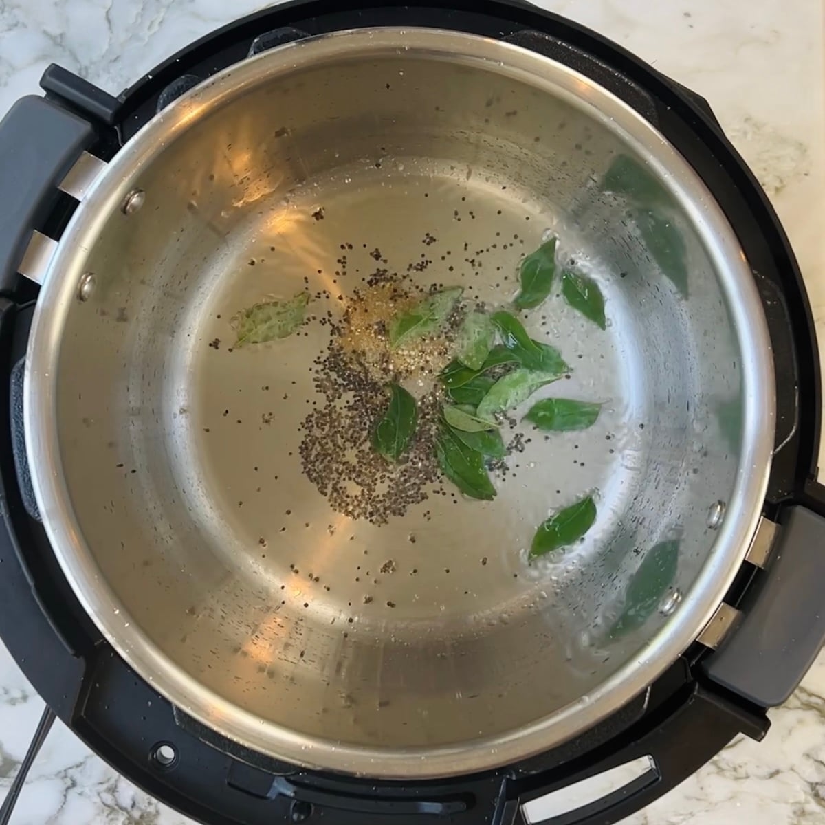 Steel pot with heated oil, mustard seeds, and fresh curry leaves inside an electric pressure cooker on a marble countertop.