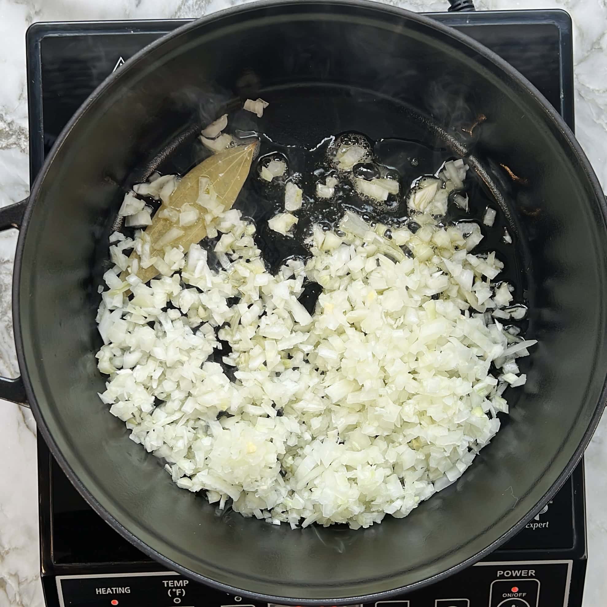 Chopped onions and a bay leaf sautéing in oil in a black pot on an electric stovetop.